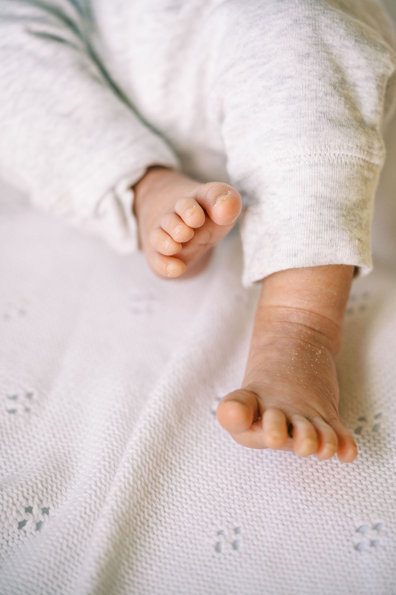 Close-up of tiny newborn baby feet resting on a white knit blanket, wearing a soft gray onesie.