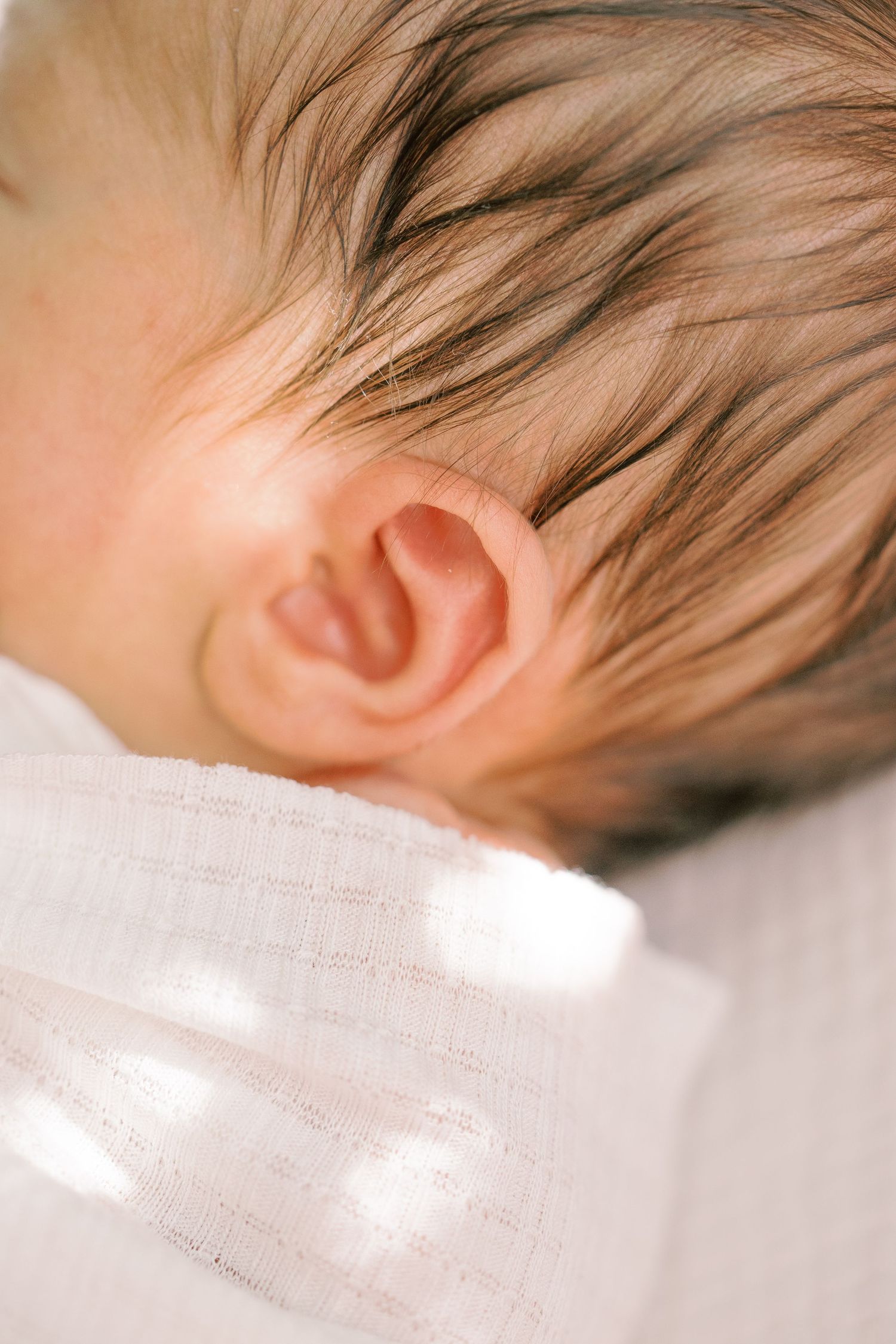 Close-up of a sleeping newborn baby's ear and fine hair resting on a soft white textured blanket.