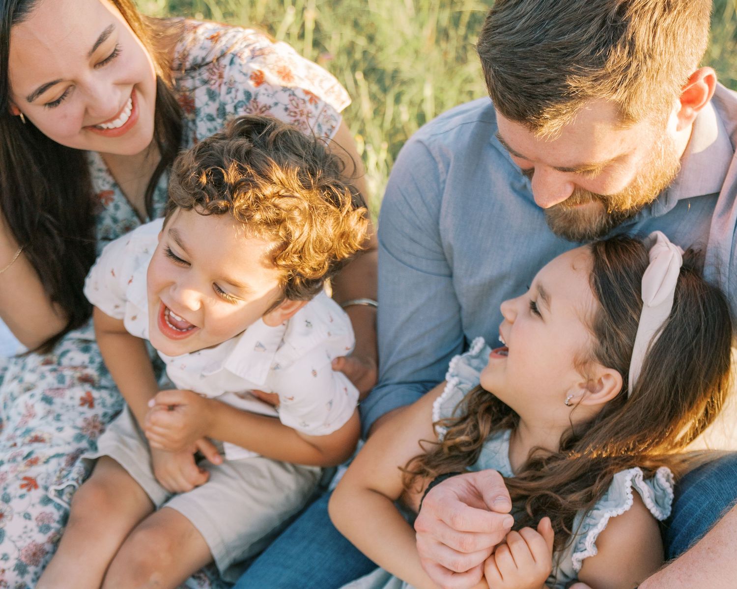 Happy family of four laughing together outdoors, parents holding two young children in a sunny field.