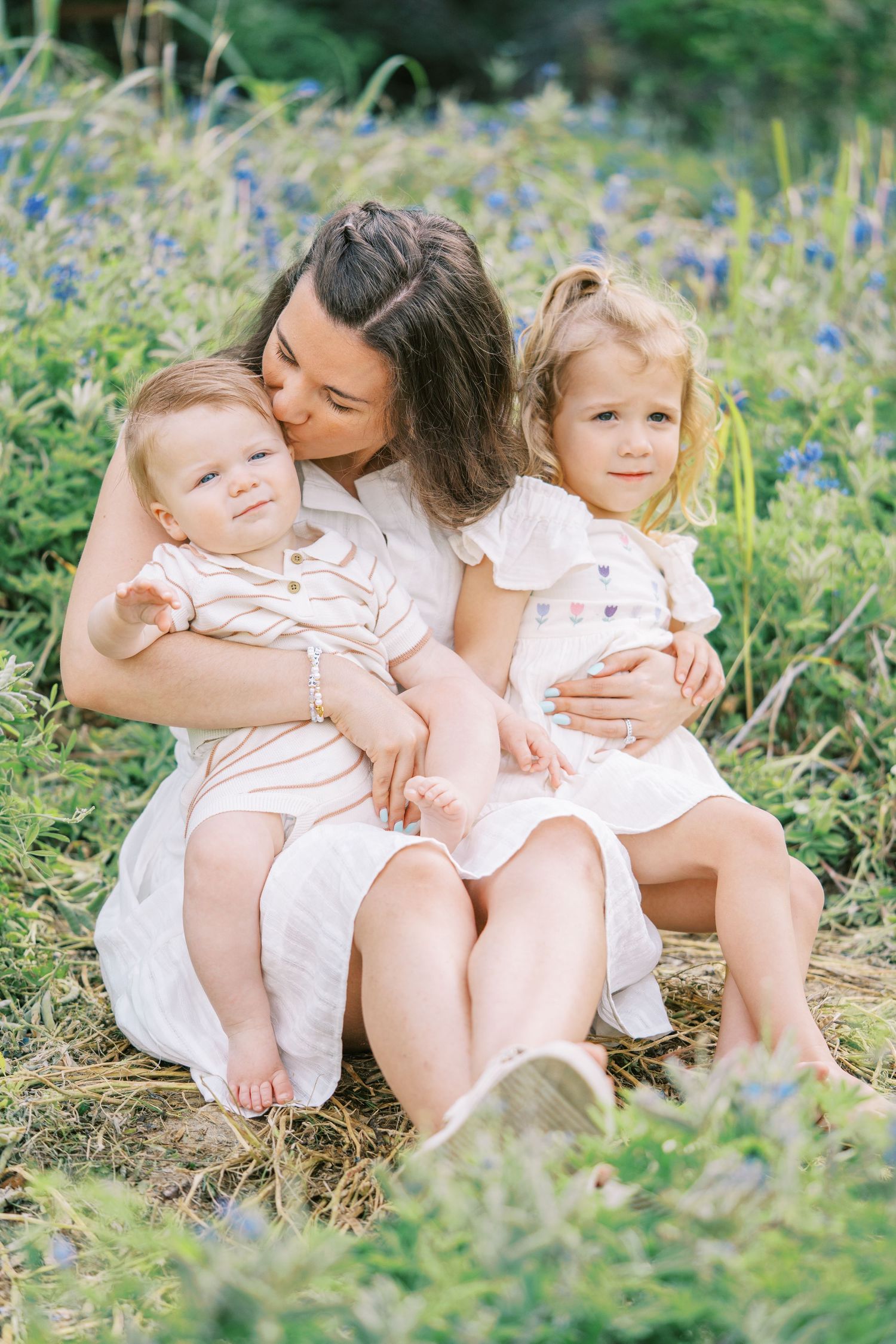 A mother sits in a wildflower field kissing her toddler while holding her baby, all dressed in white.