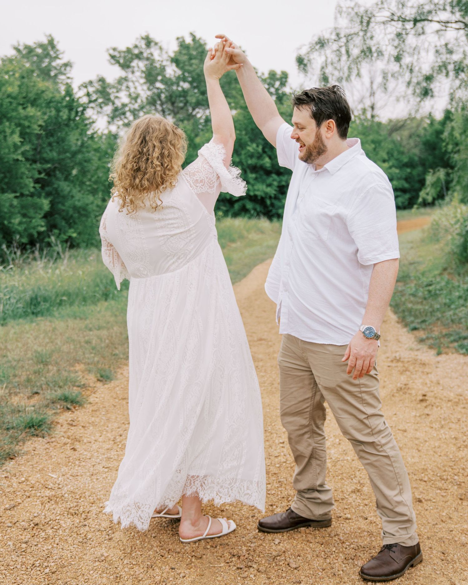 A joyful couple dances on a sunny dirt path, with the man spinning the woman in a white lace dress.