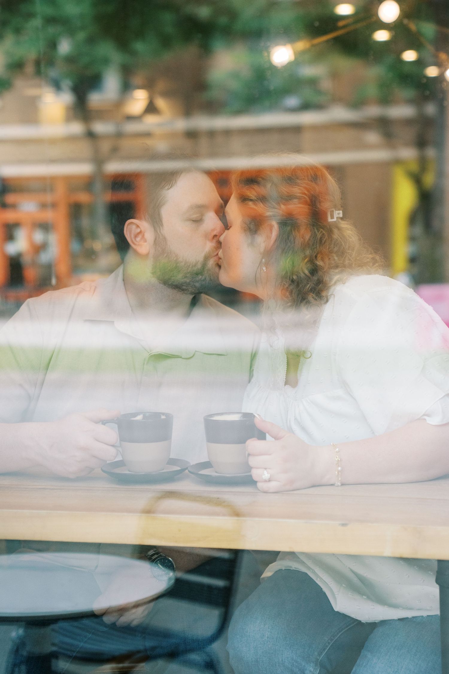 Romantic couple kissing at a cafe table with two coffee cups, photographed through a reflective window.