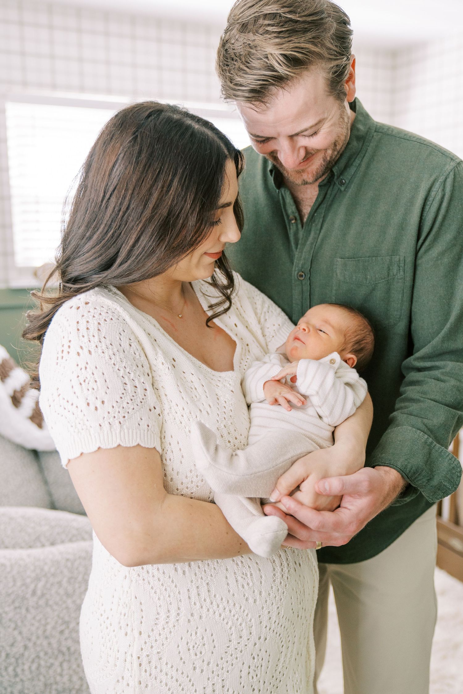 Parents in green and white outfits share tender moments with their newborn baby in a bright home setting.