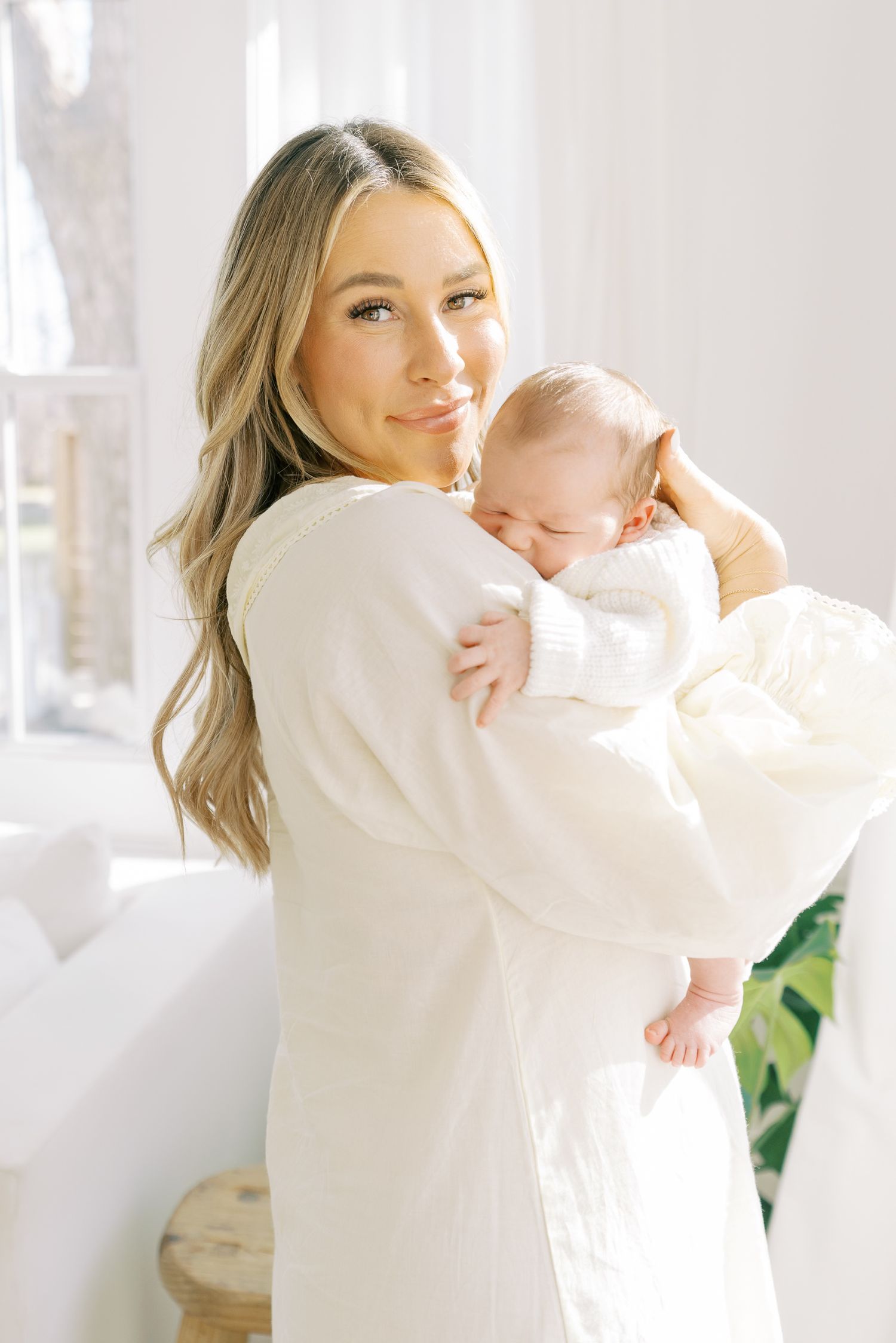 Woman in white sweater holding and embracing baby in bright sunlit room.