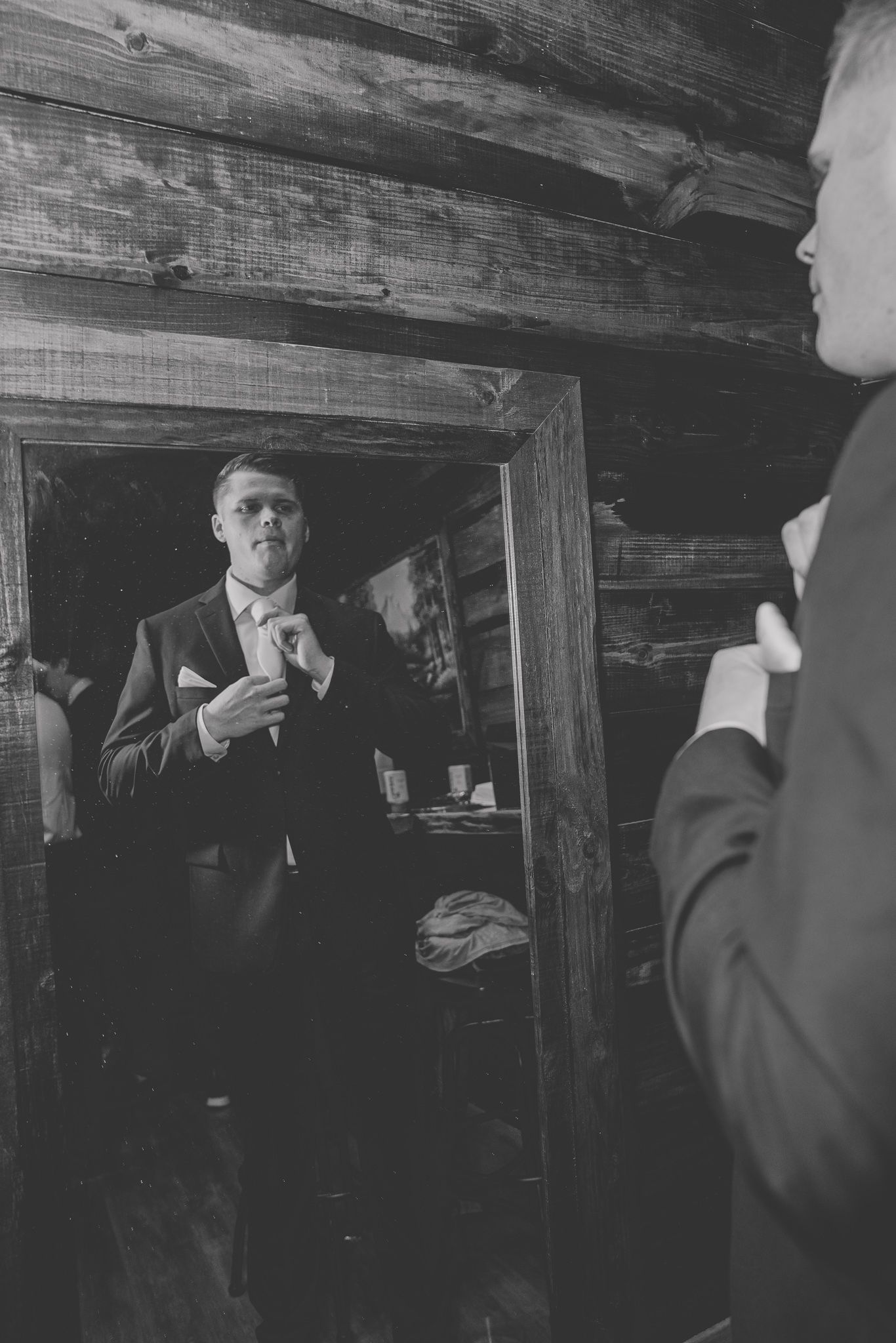 Black and white image of groom adjusting tie while looking in rustic wooden mirror.
