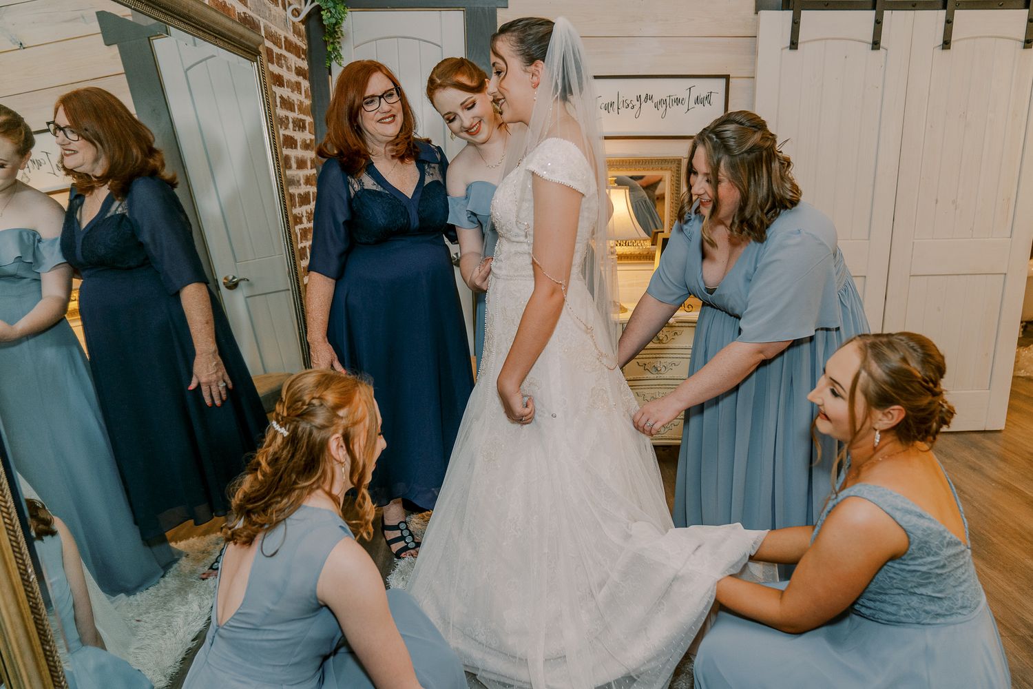 Bridesmaids in light blue dresses help adjust the train of a white wedding gown in a rustic indoor setting.