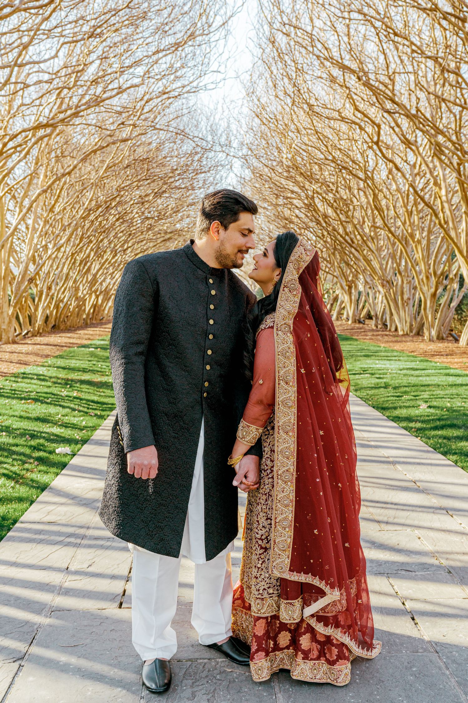 Couple walking together in traditional Indian wedding attire through a tree-lined pathway.