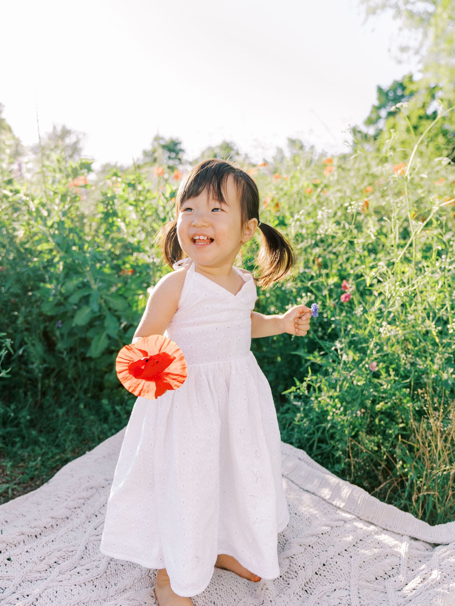 A joyful child in a white sundress dances with a red poppy flower in a garden.