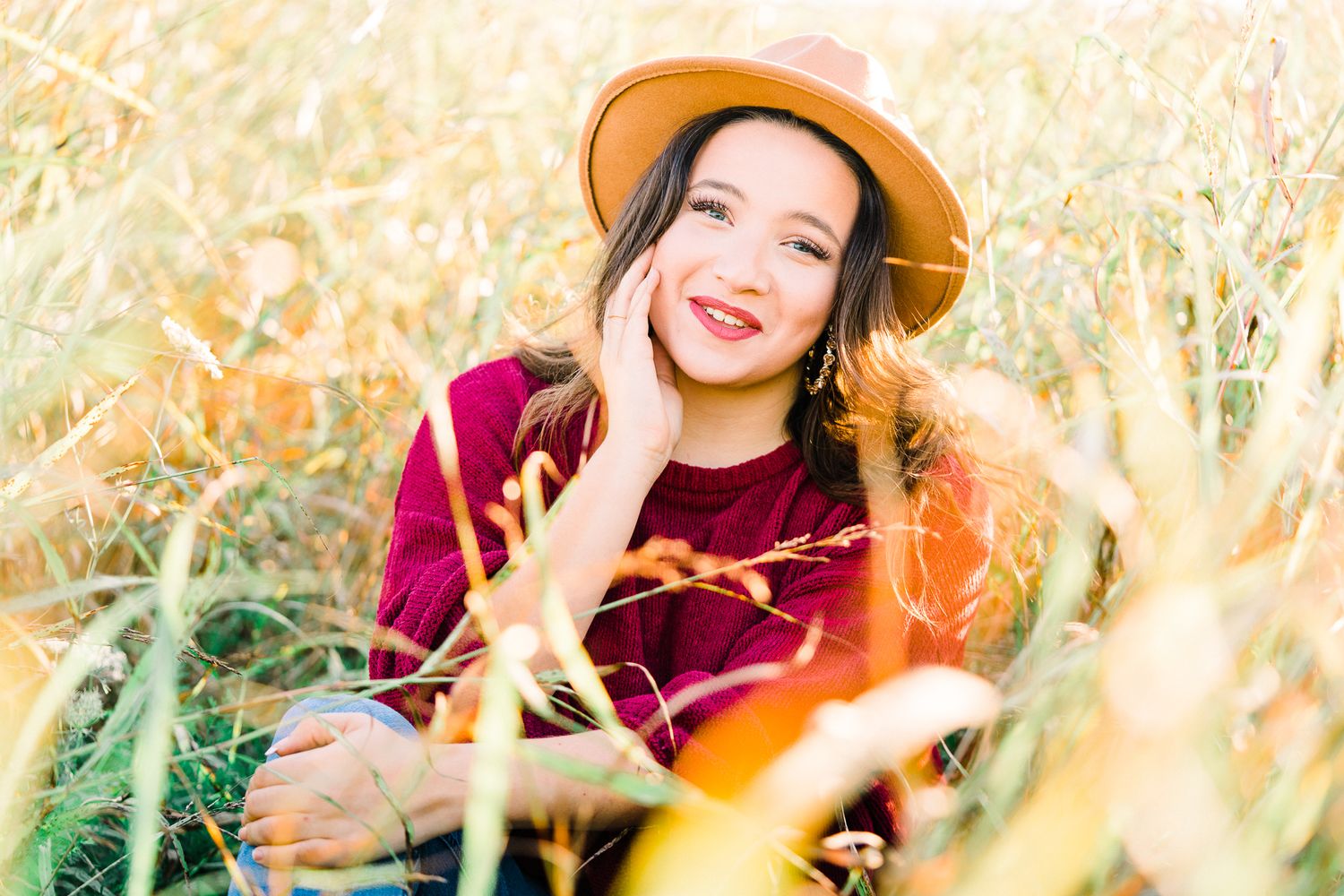 A person wearing a brown hat and maroon sweater smiles while sitting in tall golden grass during autumn.