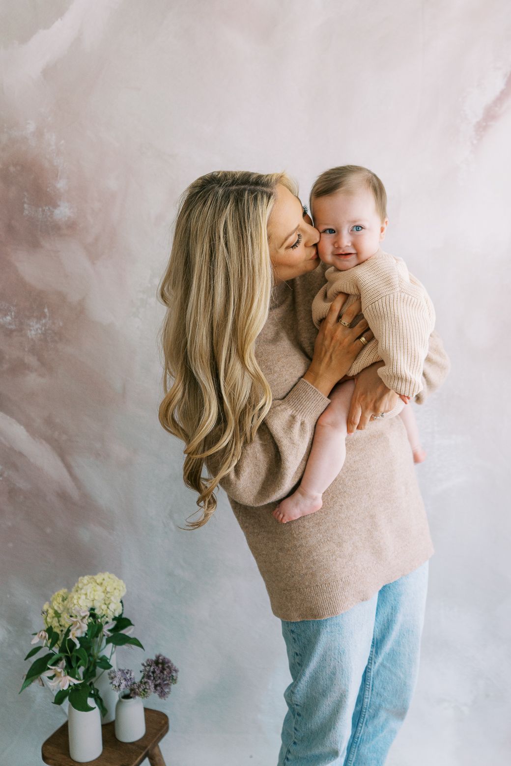 A tender moment captured between parent and baby in matching beige sweaters against a textured wall with flowers.