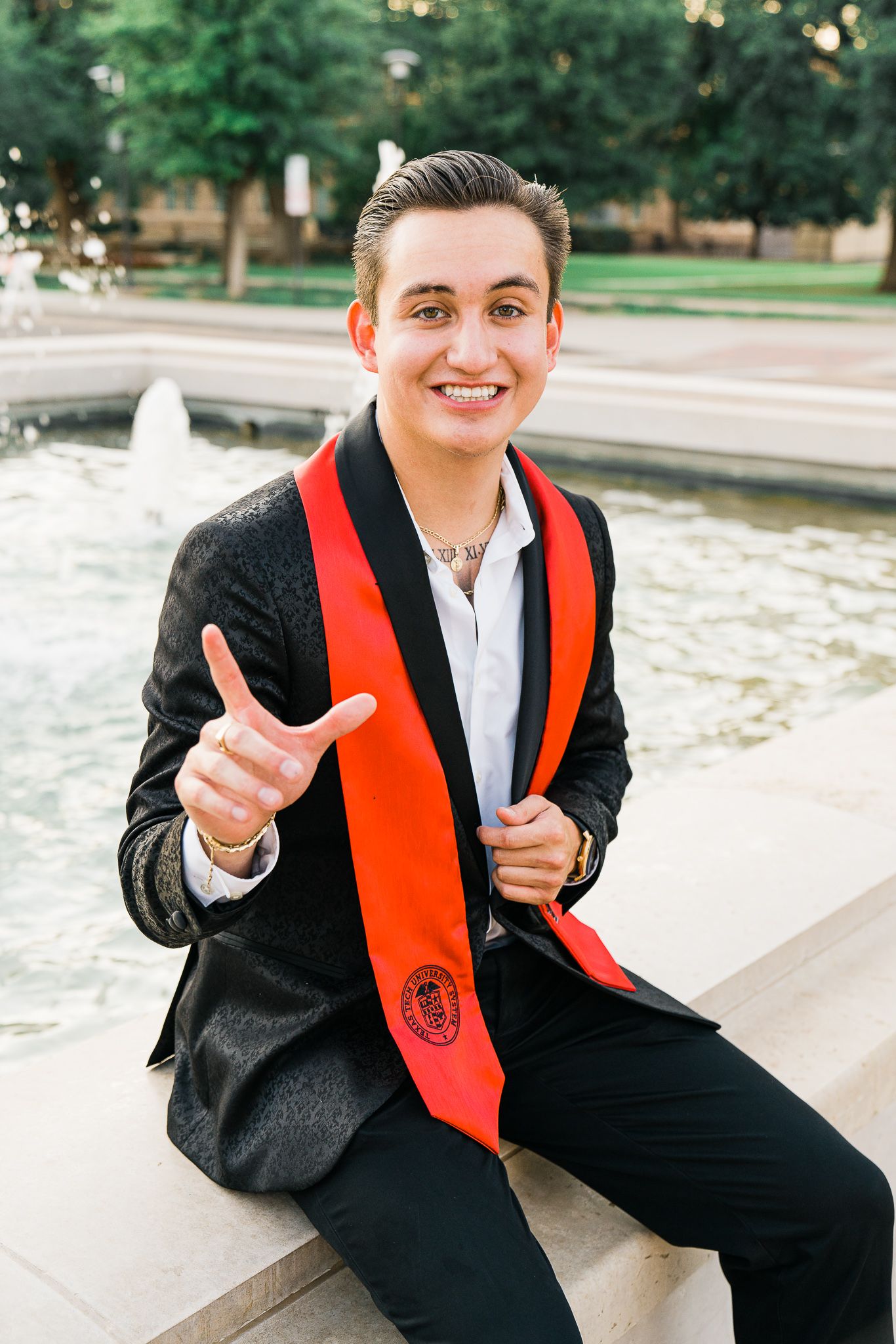 Graduate in black suit and orange stole poses outdoors with peace sign gestures.
