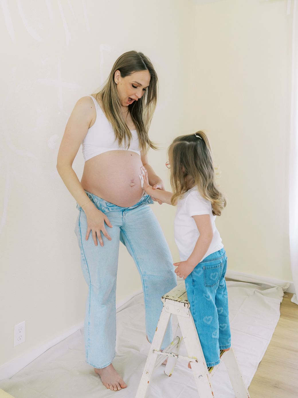 A pregnant woman and child stand together near a white stepladder in a bright room.