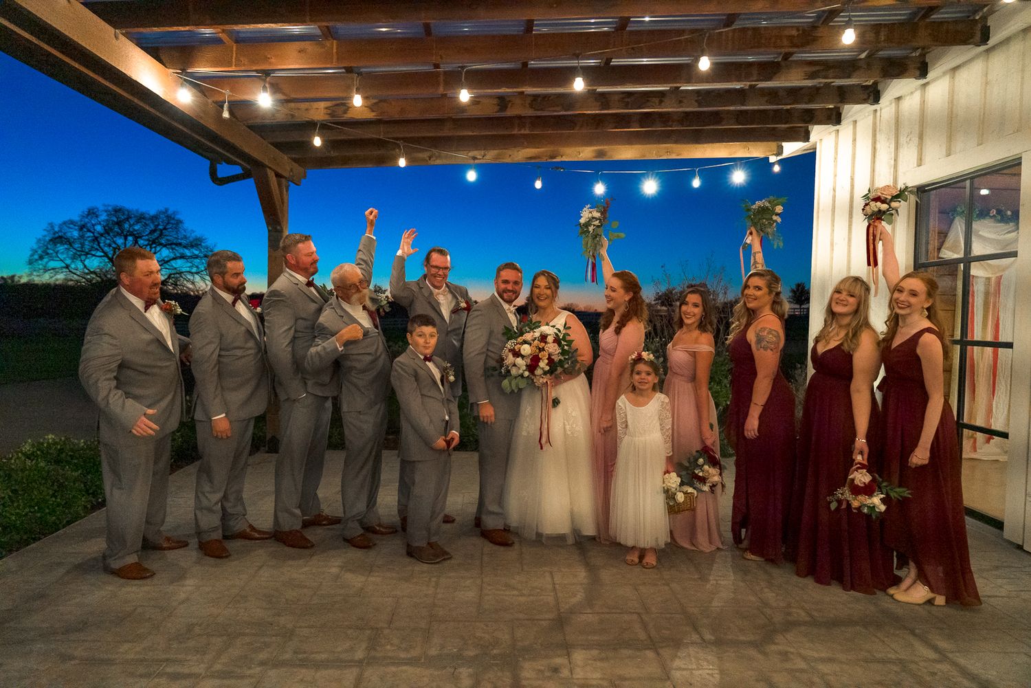 Wedding party members pose with arms raised in celebration at a rustic indoor venue with wooden beams and warm lighting.