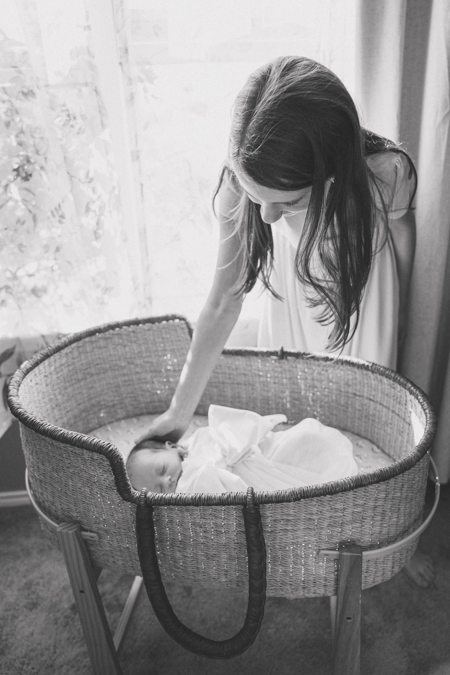 Black and white photo of a mother gently touching her newborn baby in a wicker Moses basket bassinet.