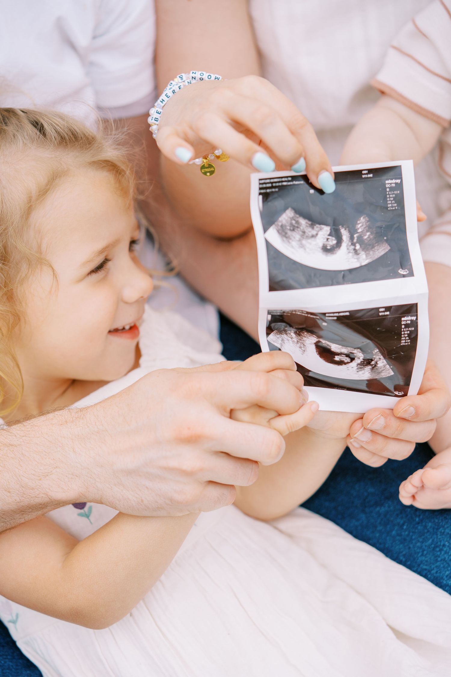A young girl in a white dress smiles while holding ultrasound photos with her pregnant mother and father.