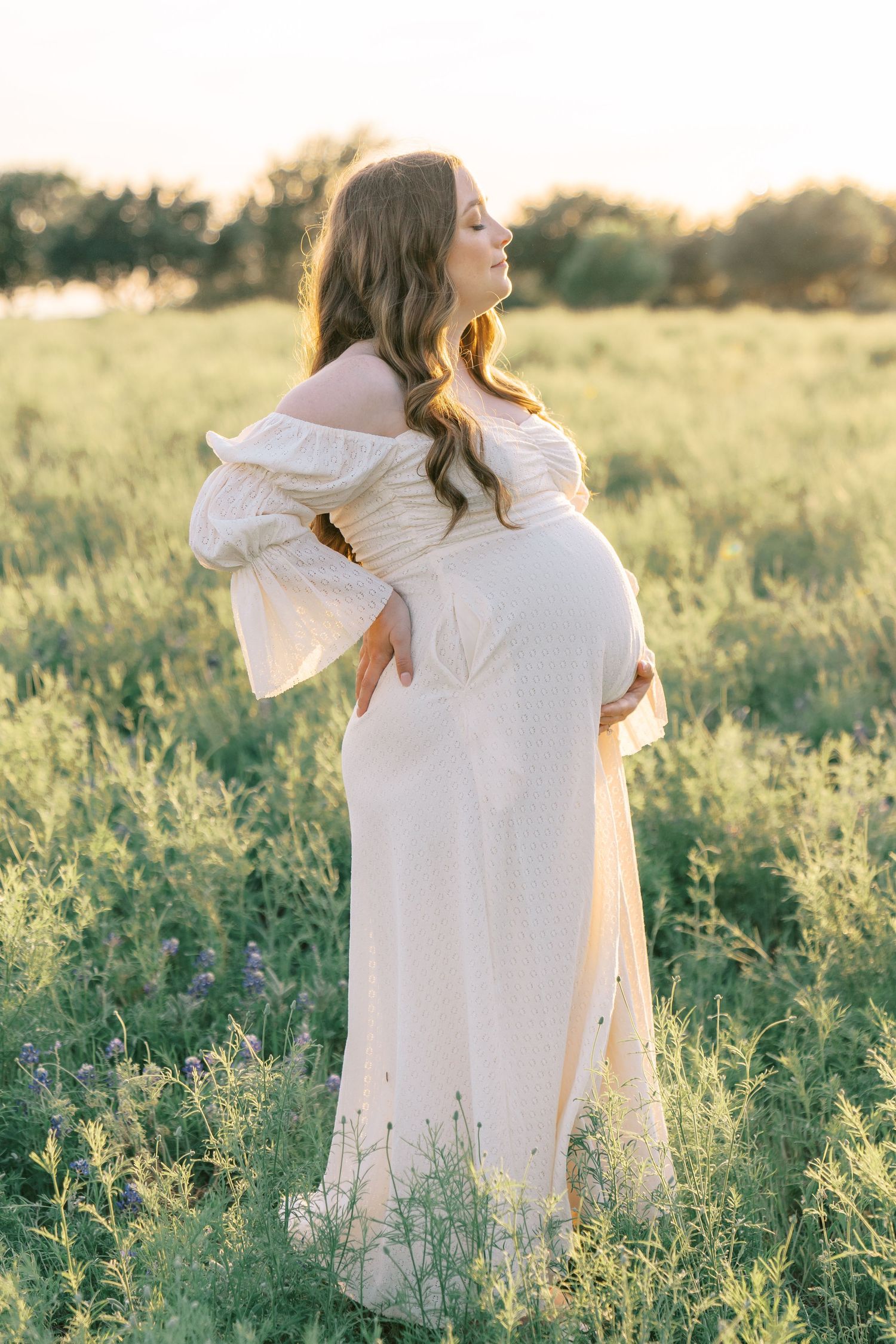 Pregnant woman in white off-shoulder gown standing in green field during golden hour sunset.