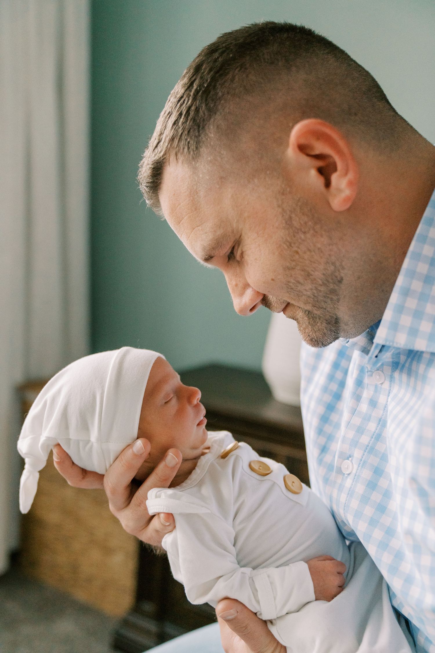 A father in a plaid shirt tenderly holds his newborn baby wearing a white outfit and knot hat.
