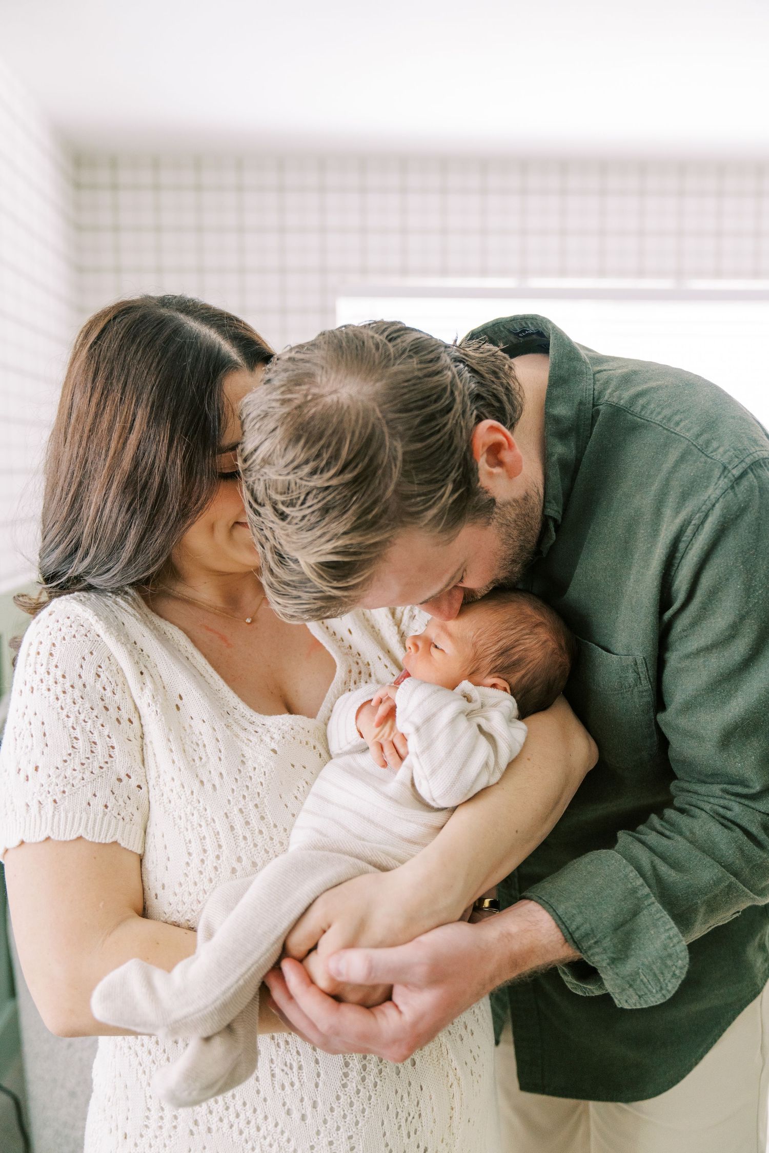 A tender family moment with parents cradling and kissing their newborn baby in a bright white room.