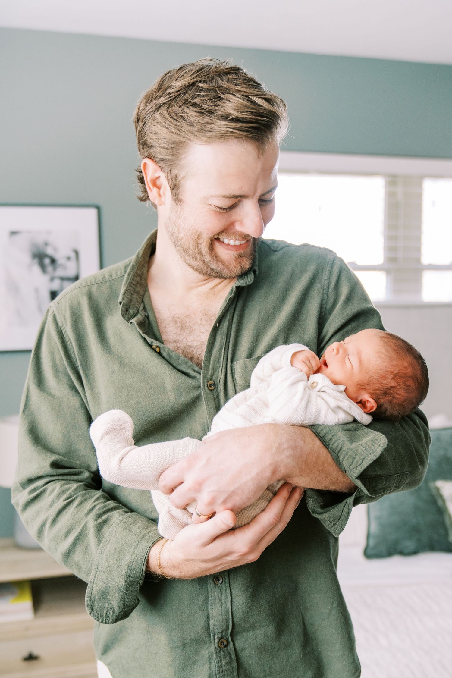 A person in a green button-down shirt smiles warmly while holding a sleeping newborn baby wrapped in white.
