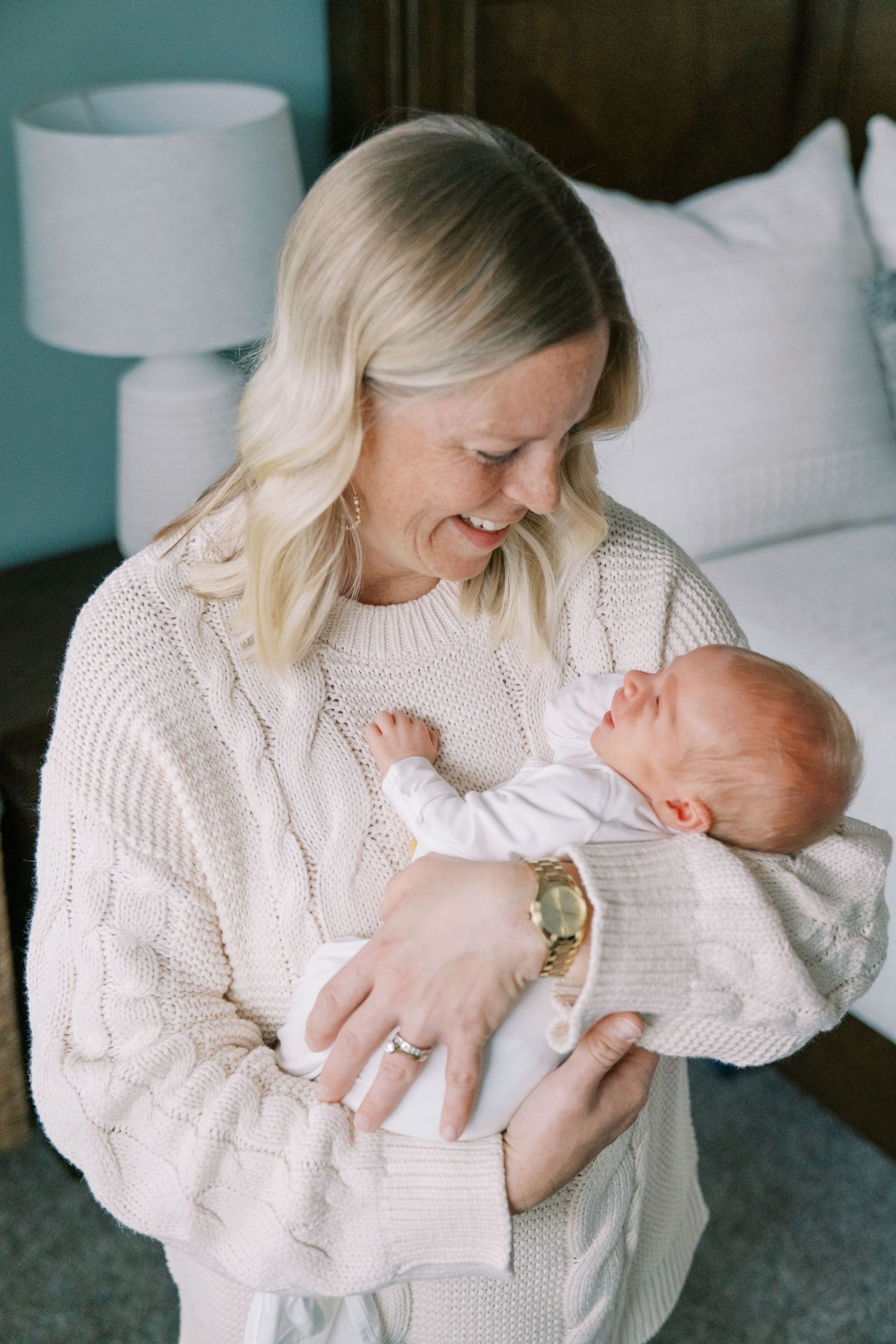 Mother in cable knit sweater snuggling with newborn baby in bedroom.