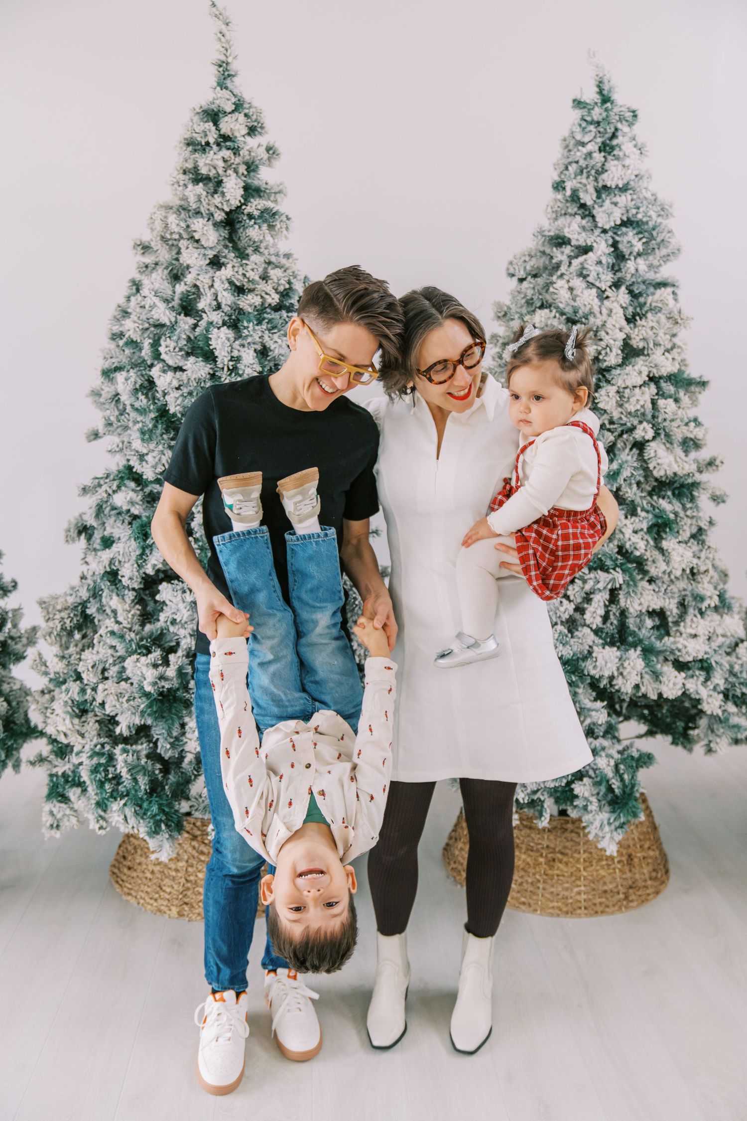 Family posing by snow-flocked Christmas trees, parents playfully holding child upside down while holding baby girl.