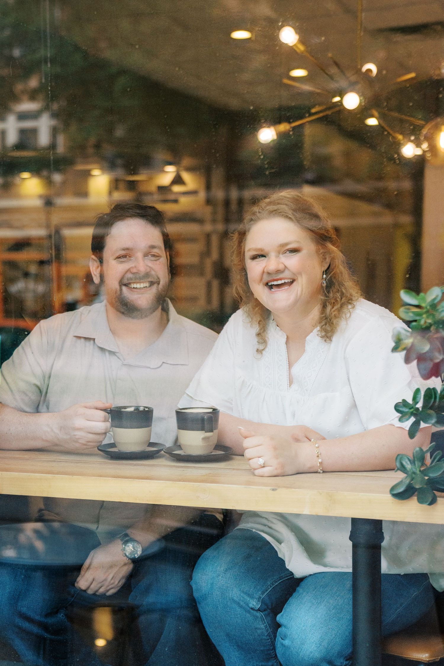 Happy couple smiling and holding coffee cups at a wooden cafe table, photographed through a window with warm string lights.