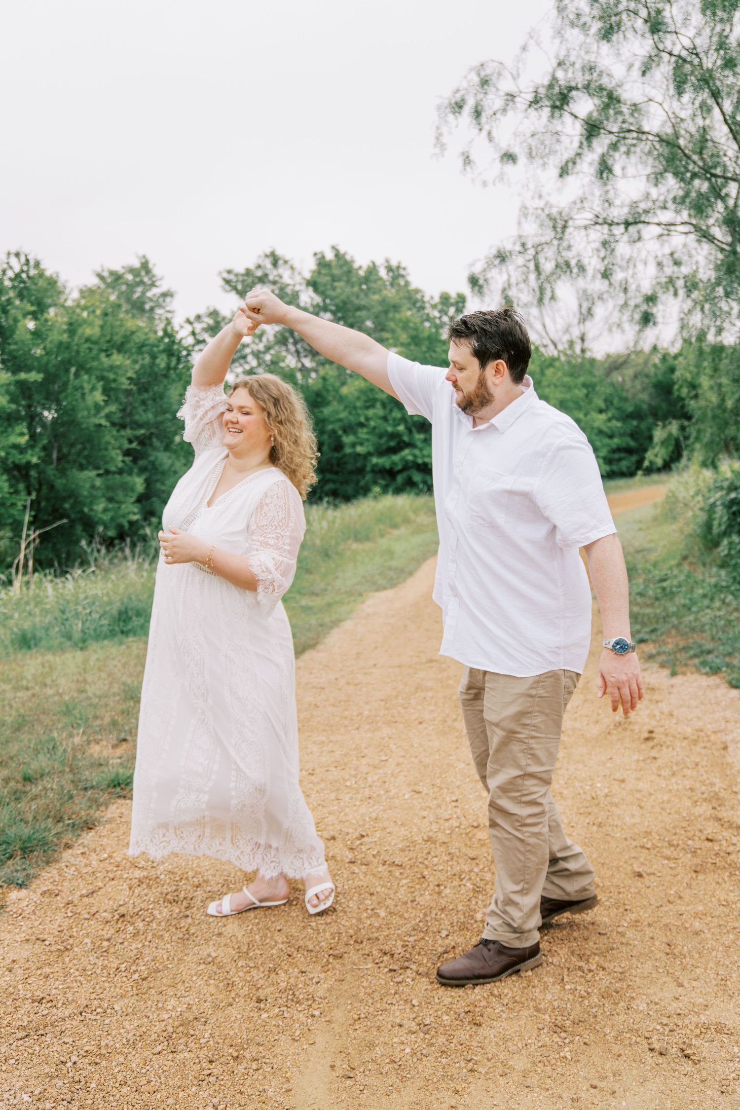 Groom spinning bride in white lace dress on a dirt path surrounded by lush greenery.