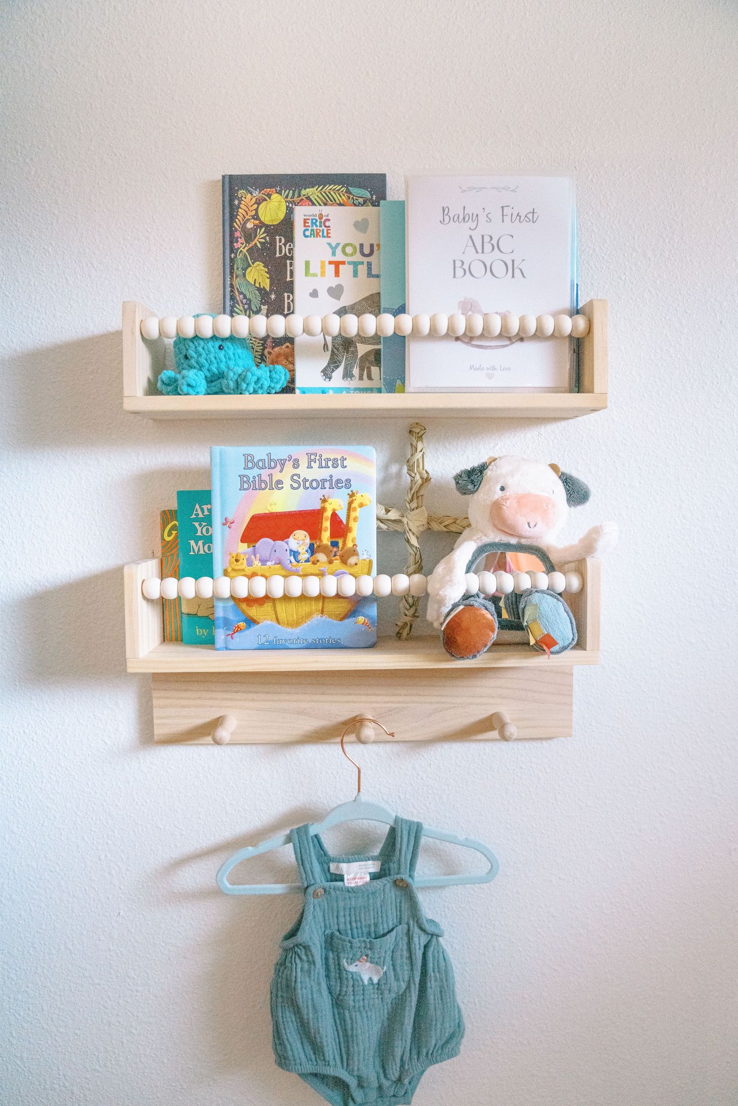 Two wooden bead shelves mounted on wall displaying baby books, stuffed animals, and a teal romper hanging below.