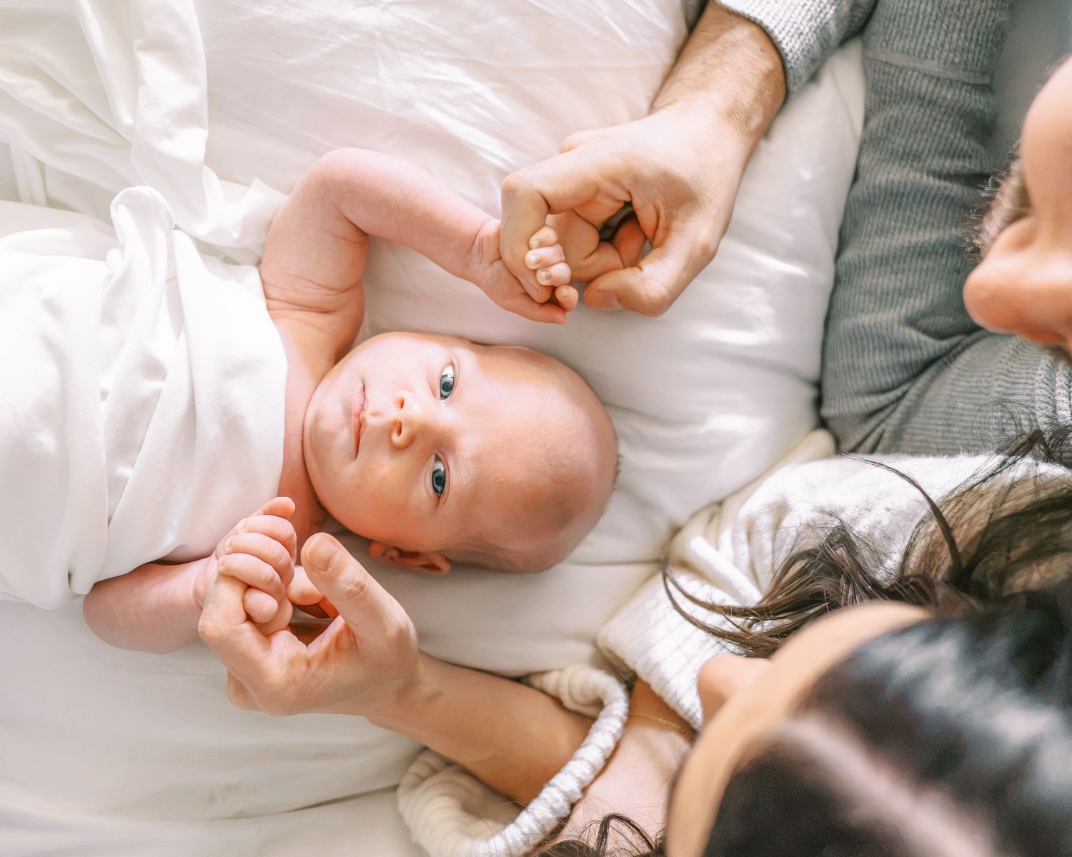 A newborn baby lying on a white bed between two parents, looking up while holding their fingers.
