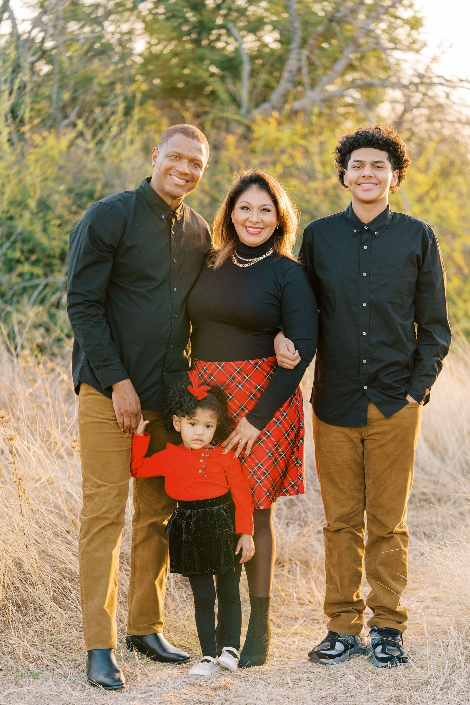 A family in coordinated black and red outfits poses together in a golden field during sunset.