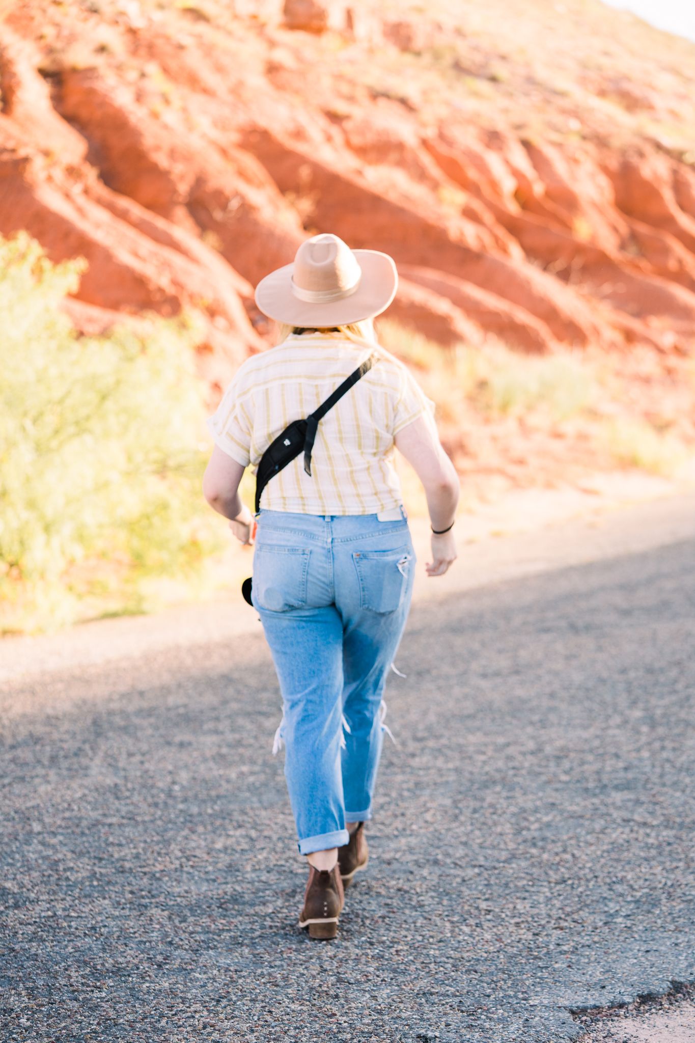 A person in casual attire walks along a desert road with red cliffs in the background.