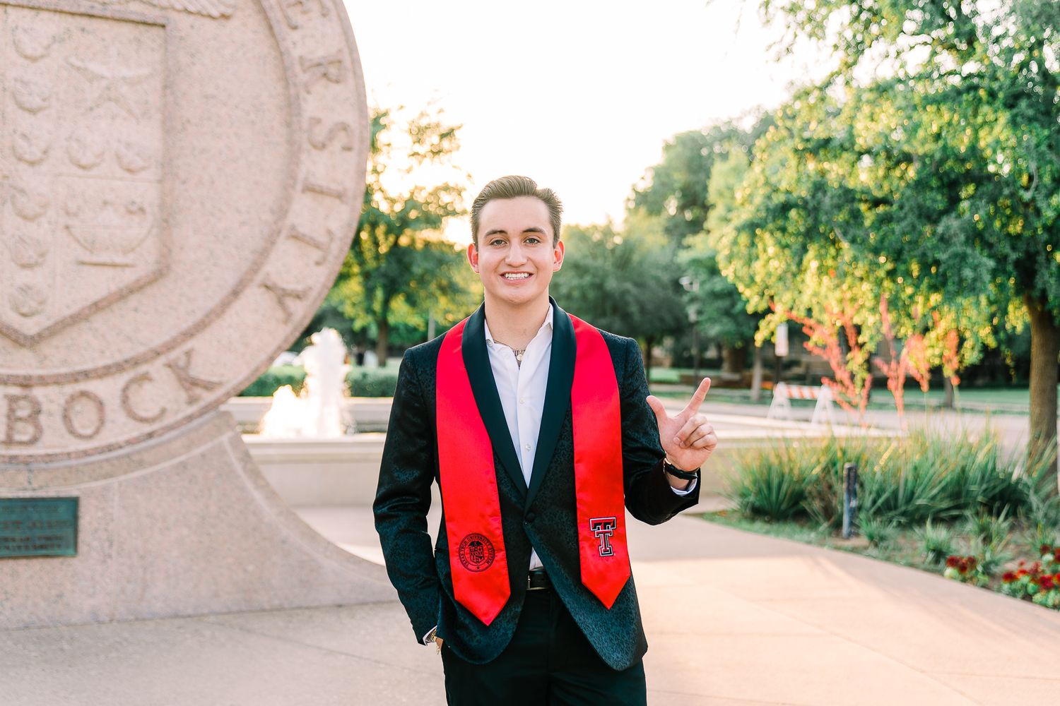 A graduate wearing a black suit and red graduation stole stands smiling in front of a stone campus monument.