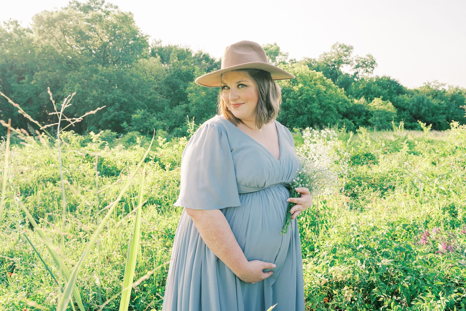 Woman in blue dress and sun hat walking through a lush green field on a bright sunny day.