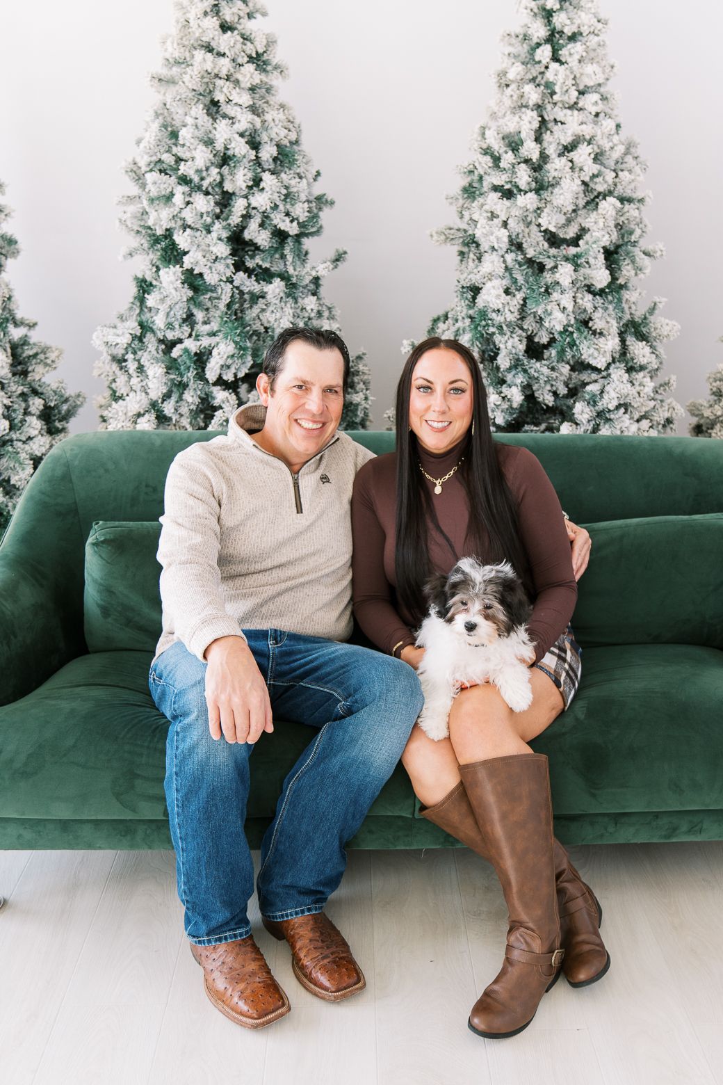 A couple sits with their small dog on a green velvet couch with snow-covered Christmas trees in the background.