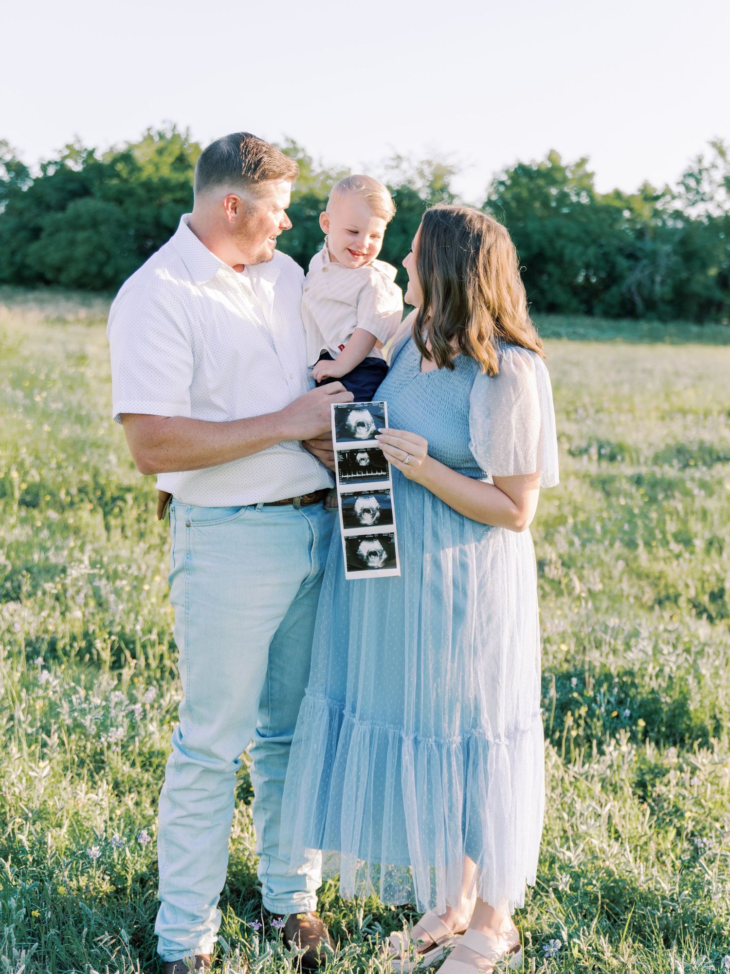 Family pregnancy announcement photoshoot in a sunny meadow with ultrasound photos and light blue dress.