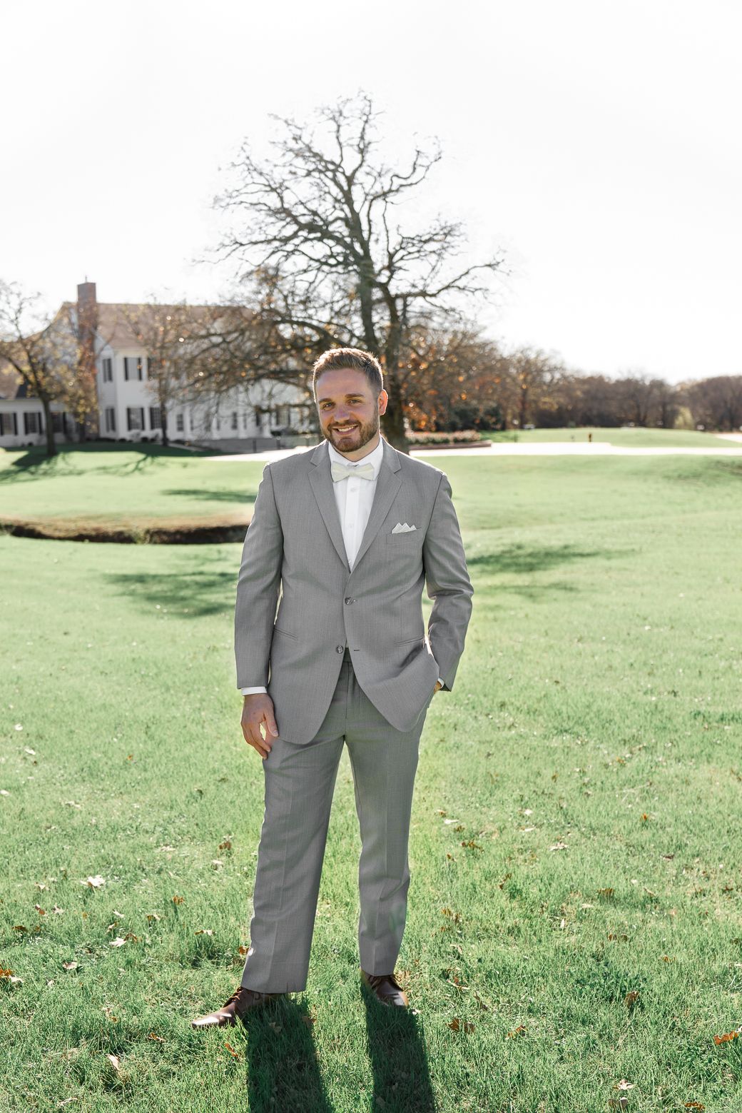 A person in a light gray suit stands on a grassy lawn with trees and a building visible in the background.