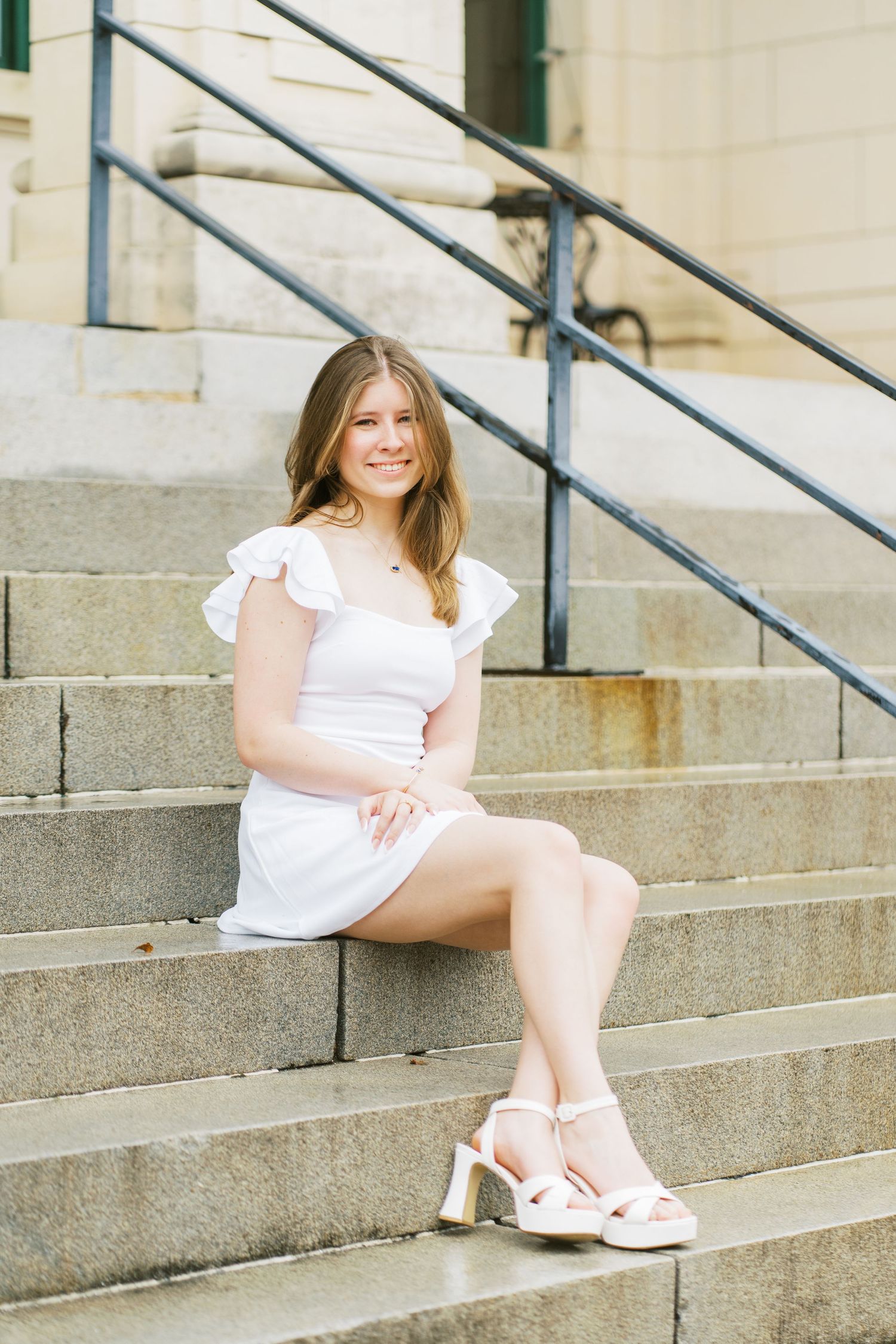 Woman in white dress sits casually on concrete steps in a bright outdoor setting.