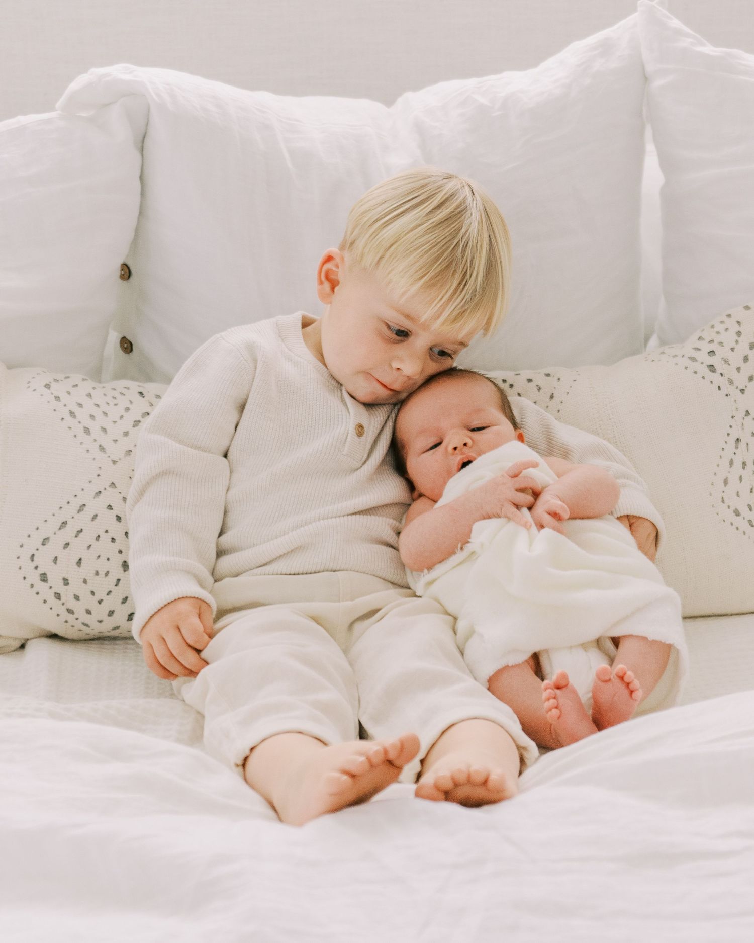 Two siblings sharing a tender moment while lying on white bedding.