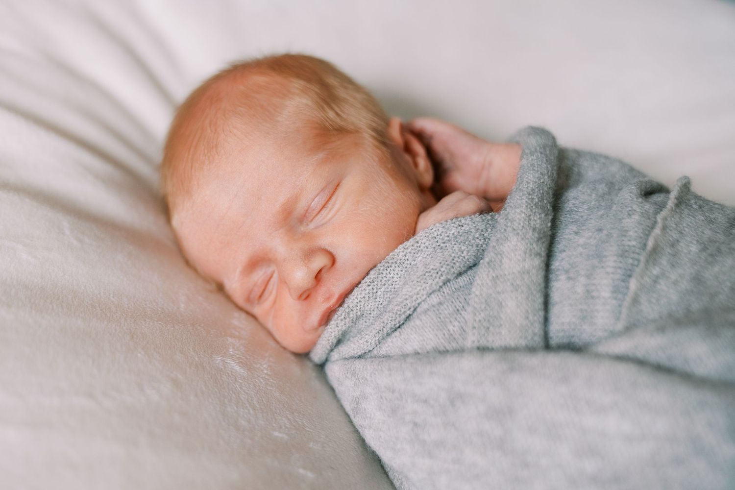 Sleeping newborn wrapped in soft gray blanket on white bedding.