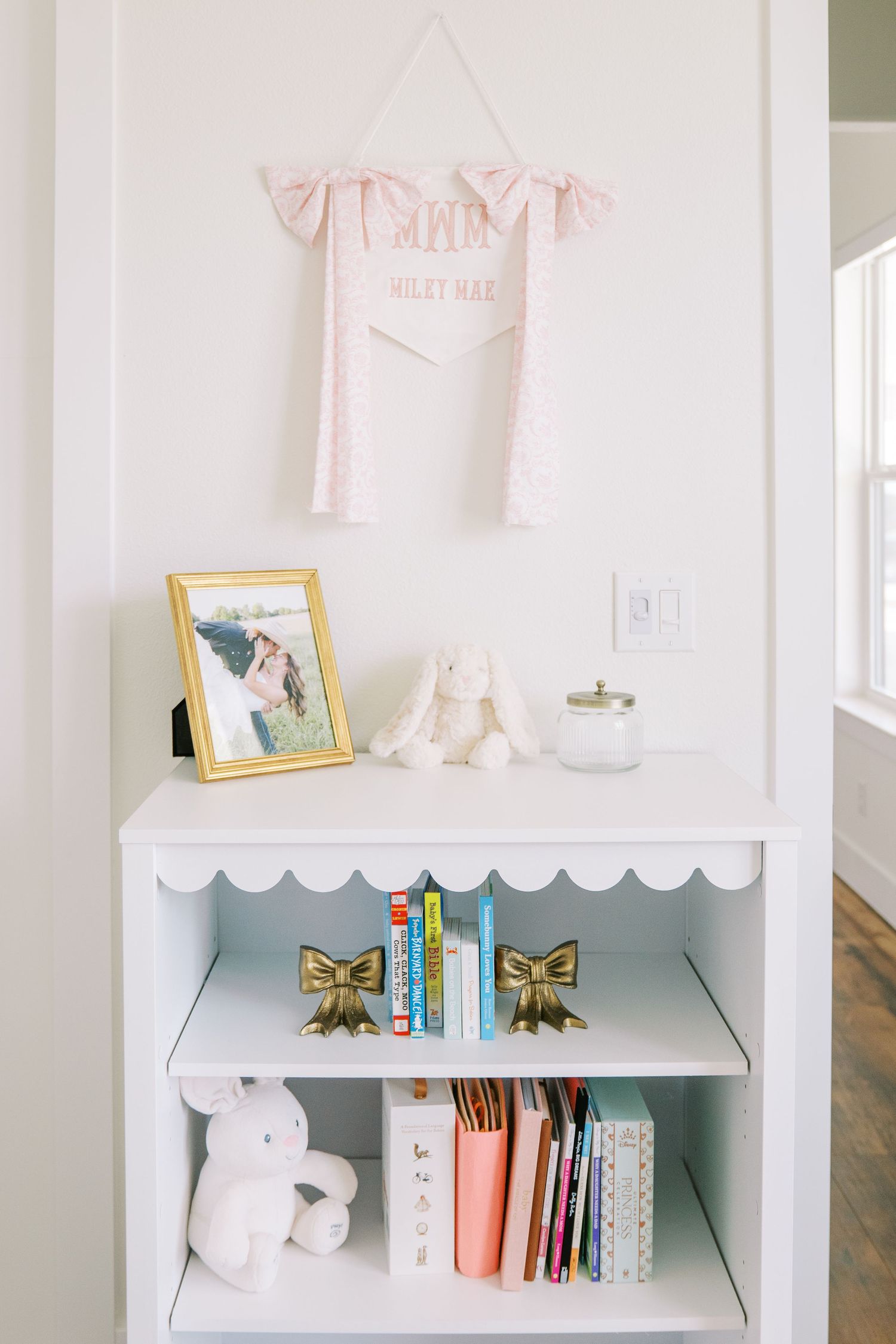 White scalloped bookshelf with gold bow bookends, children's books, stuffed animals, and a framed photo in a bright nursery.