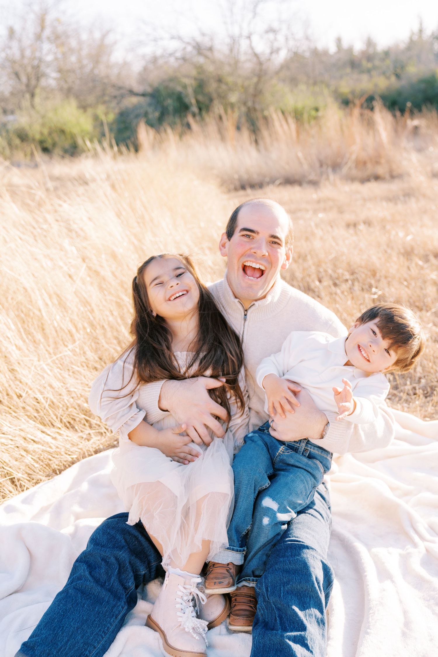 Family laughing and playing together on white blanket in tall grass field.