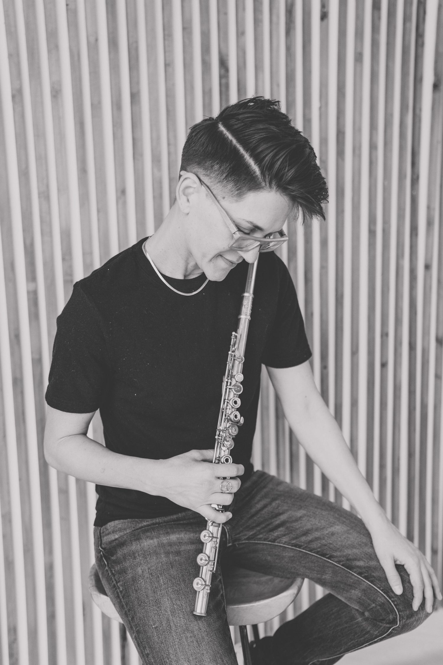 Black and white portrait of a musician playing flute while seated against a striped wall.