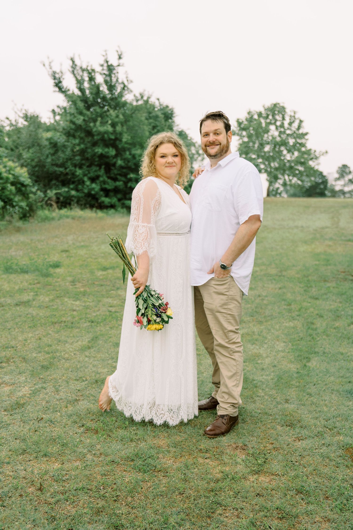 Smiling engaged couple posing in green field, woman in lace bridal gown holding colorful bouquet.