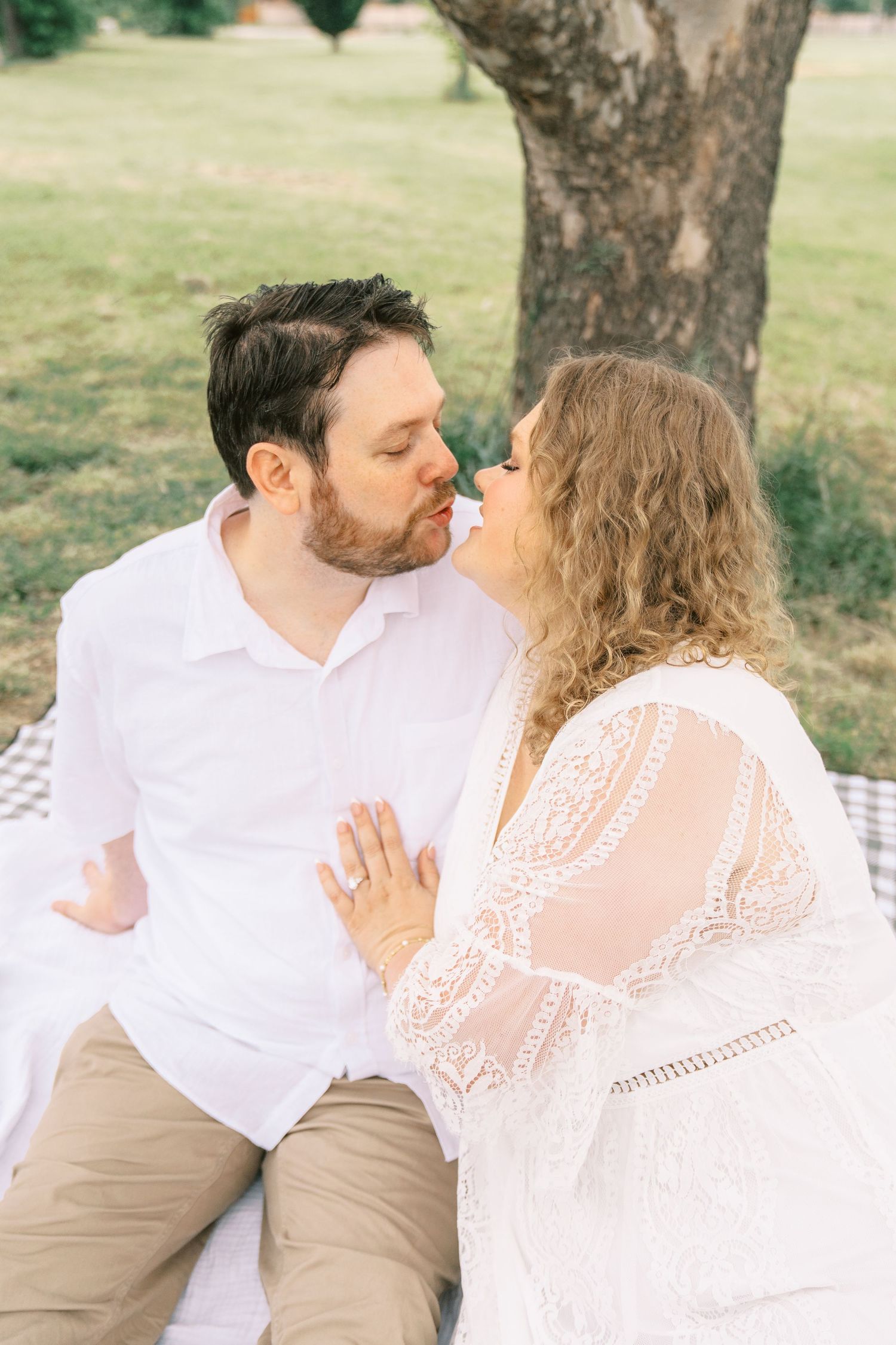 Couple sharing a romantic almost-kiss while sitting on a picnic blanket outdoors, woman wearing lace dress and engagement ring.