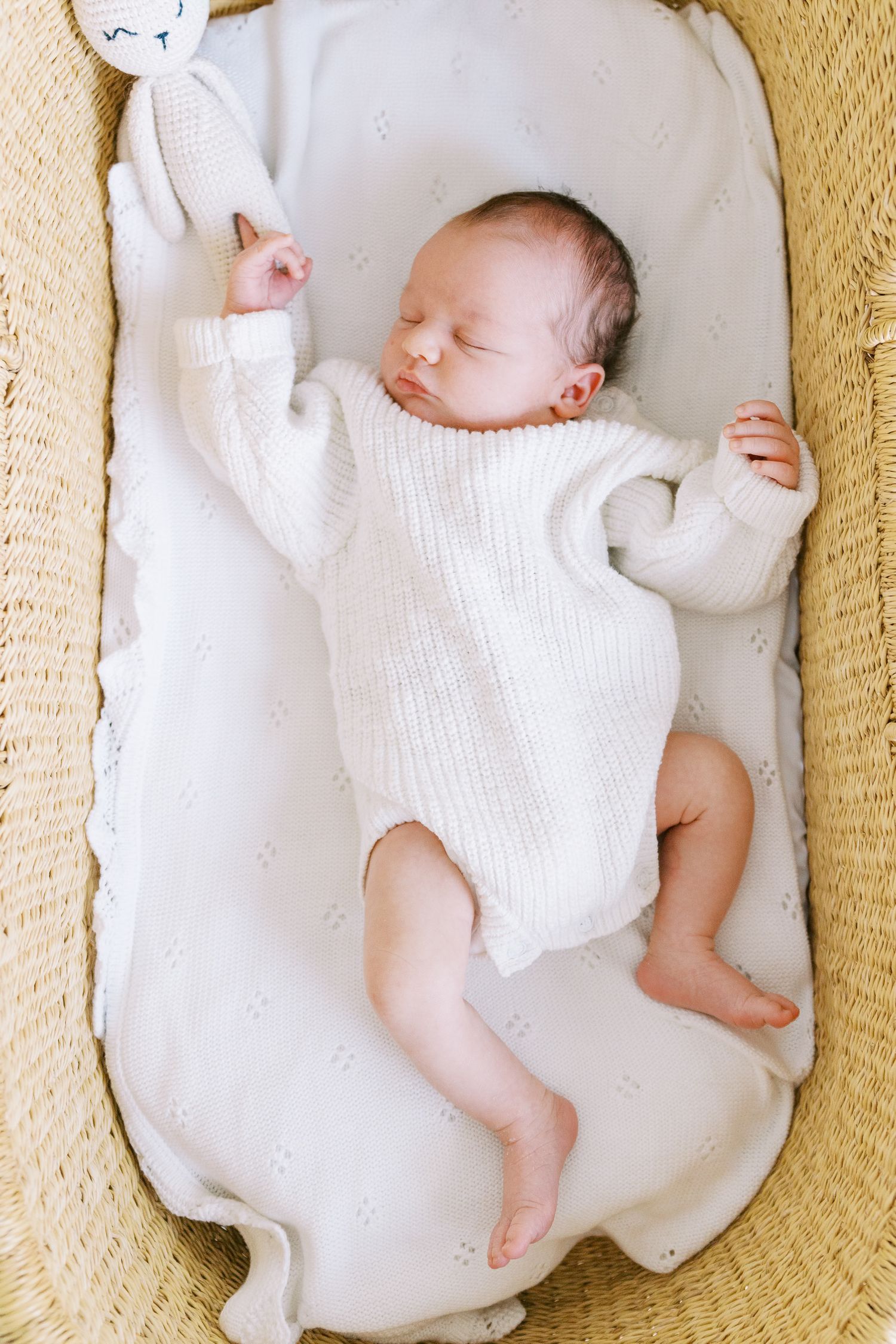 Sleeping newborn in white knit onesie resting on cream-colored cushion.