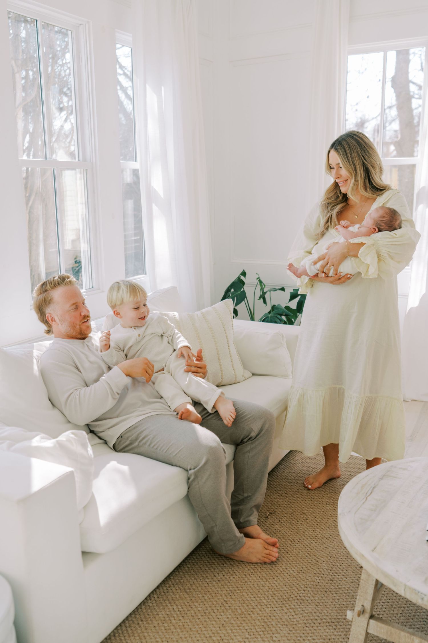 Family relaxing together in bright white living room with natural light streaming through windows.