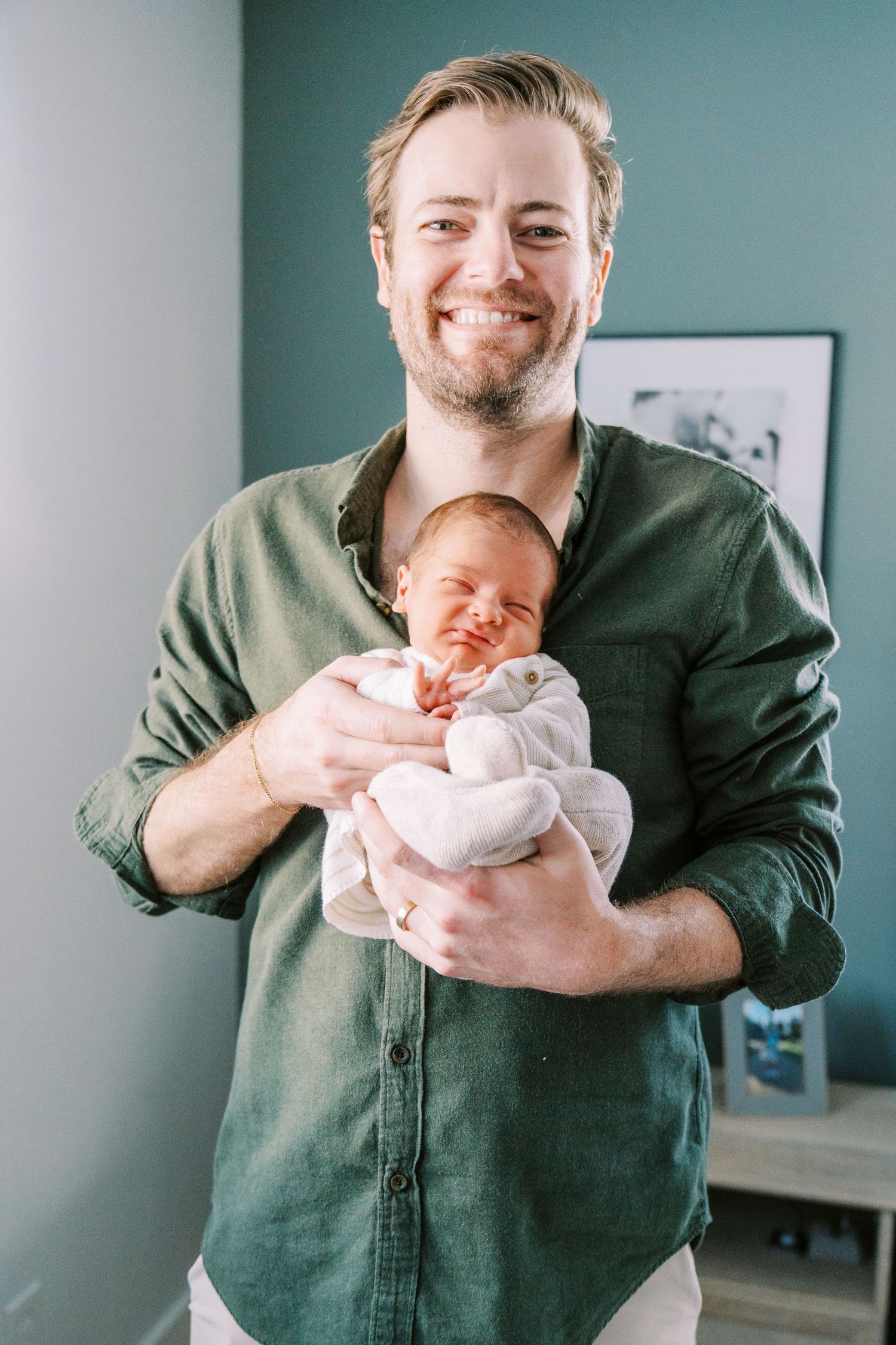 A person in a green button-down shirt holds a sleeping newborn baby wrapped in white blankets against a teal wall.