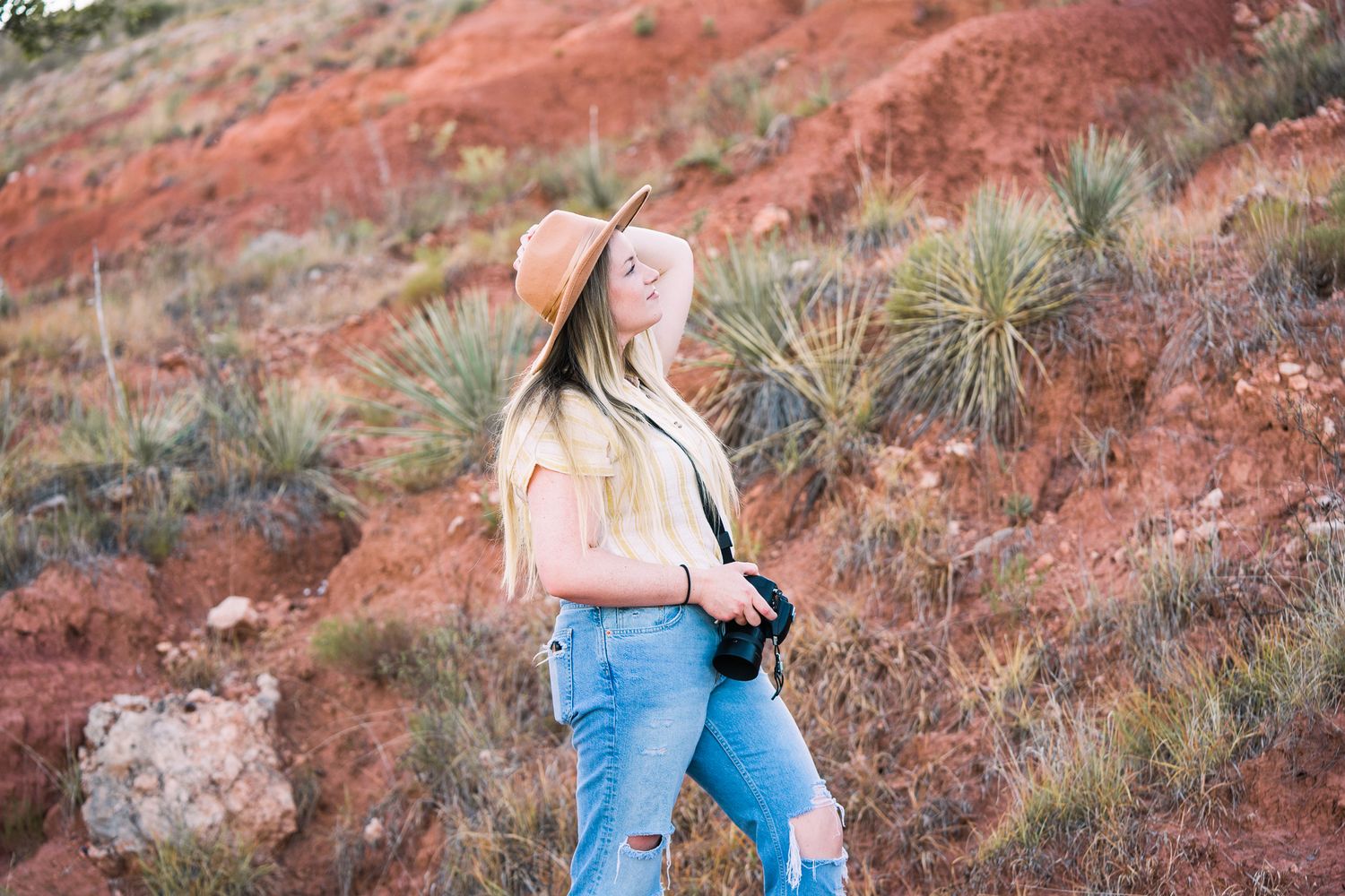 Photographer crouches with camera equipment against backdrop of red rock formations and desert plants.