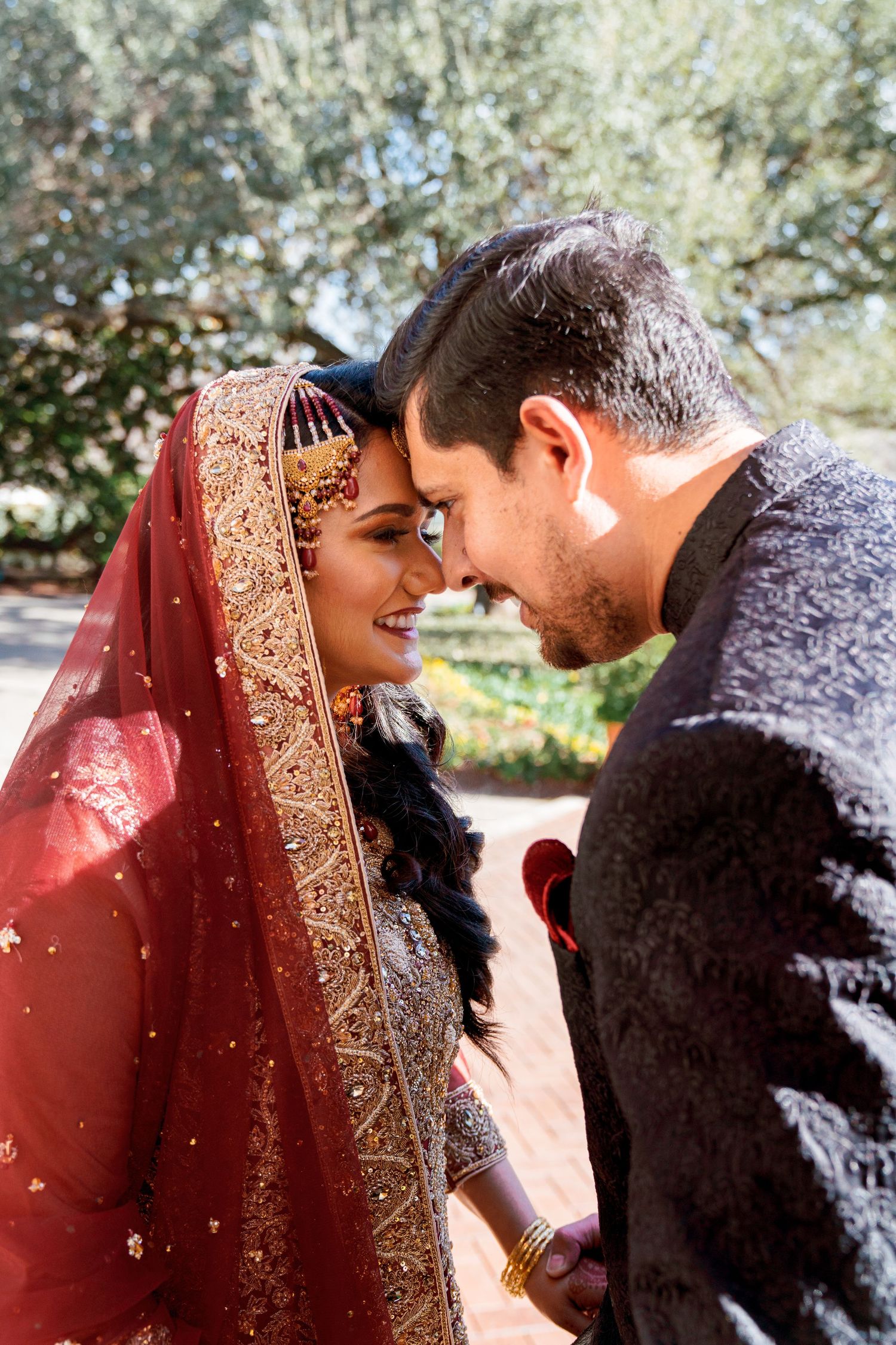 A romantic couple shares an intimate moment outdoors with the bride wearing a traditional red and gold embroidered lehenga.