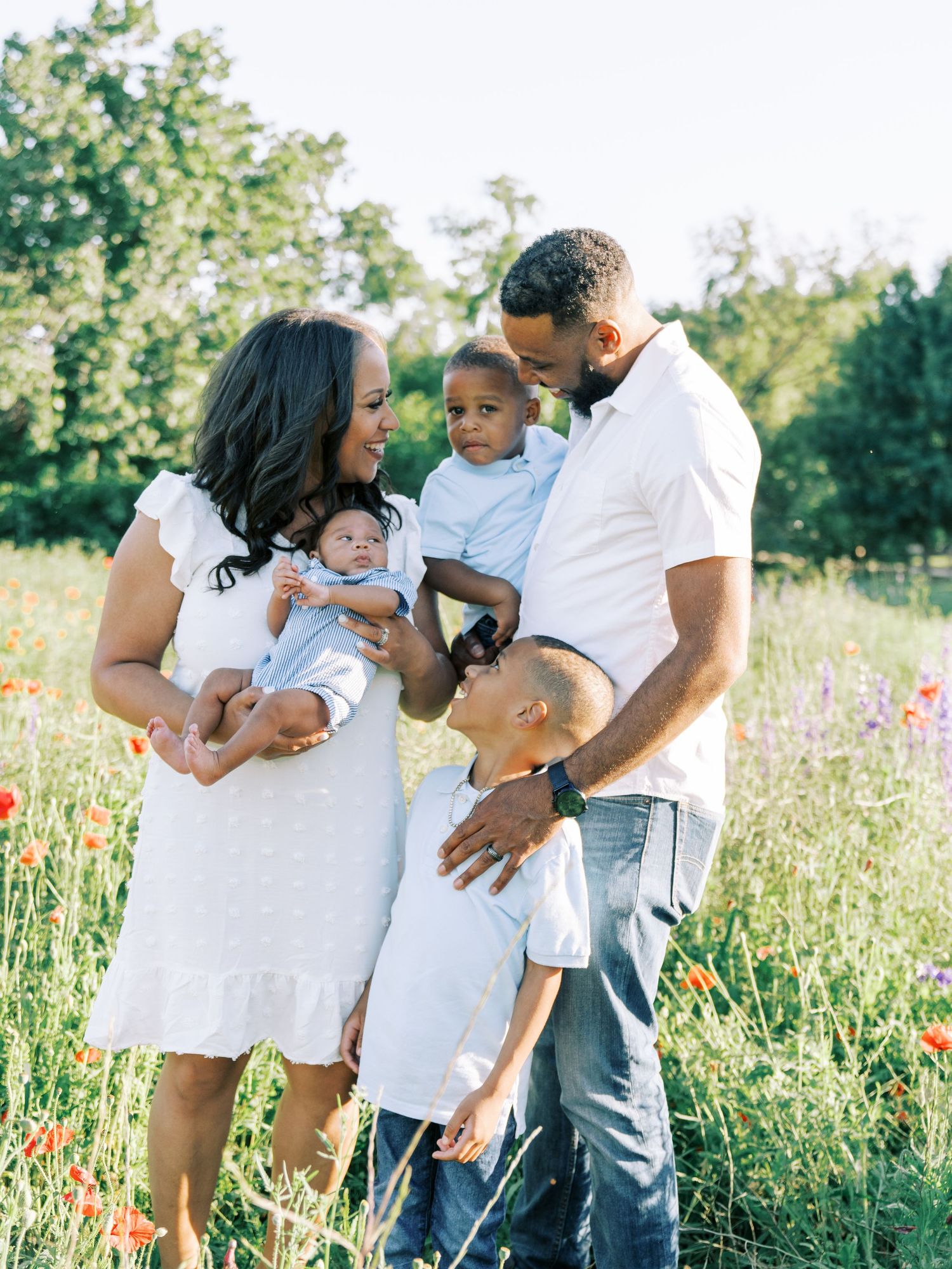 Family wearing white outfits embraces and plays together in a sunlit wildflower meadow during golden hour.